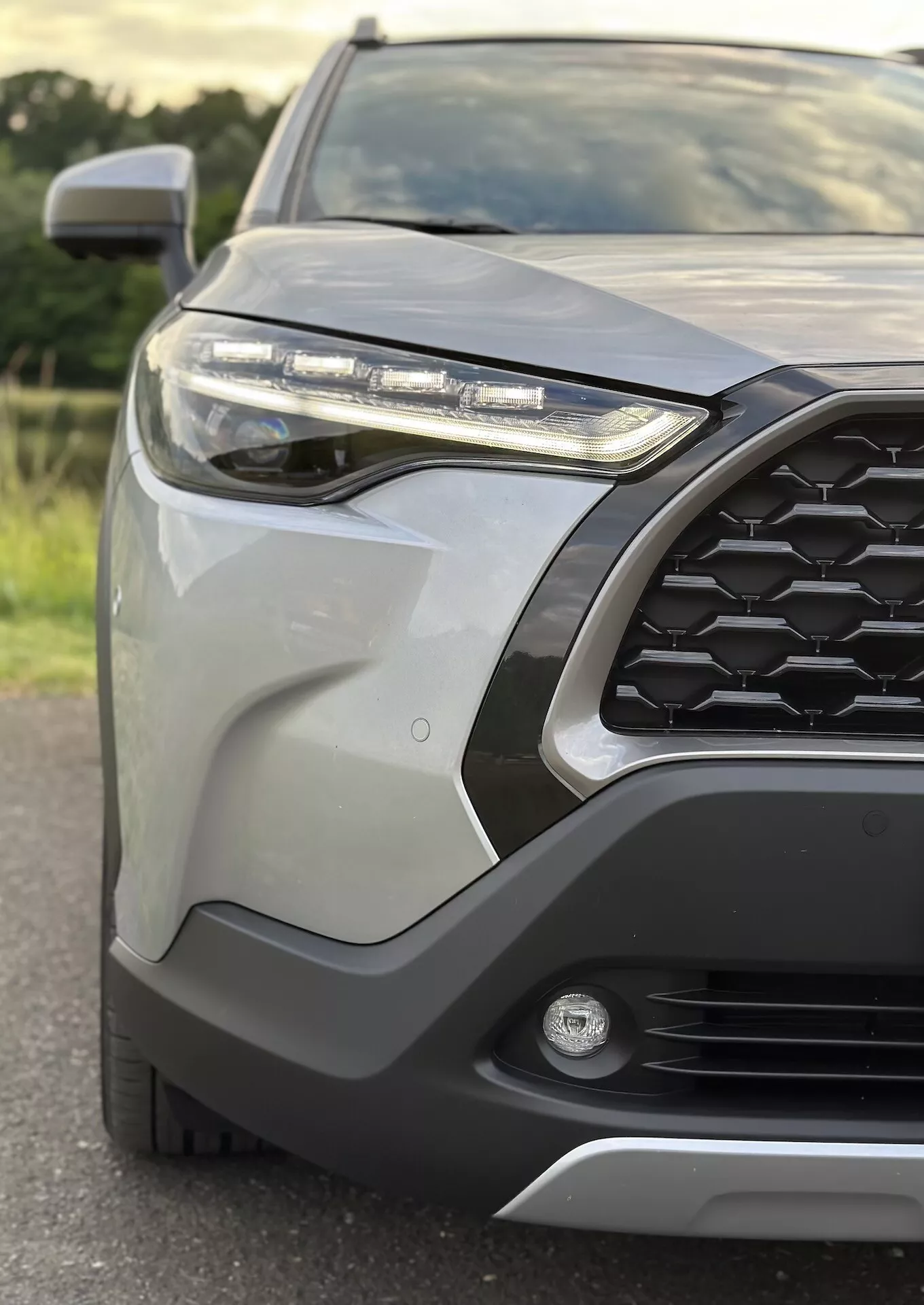 Detailní záběr na přední levou část Toyota Corolla Cross 2022 s vyobrazením vzoru šestiúhelníkové mřížky, LED světlometu, mlhového světla, předního nárazníku s ochranným krytem, elegantních karosářských dílů, bočního zrcátka s integrovaným směrovým signálem a metalického laku