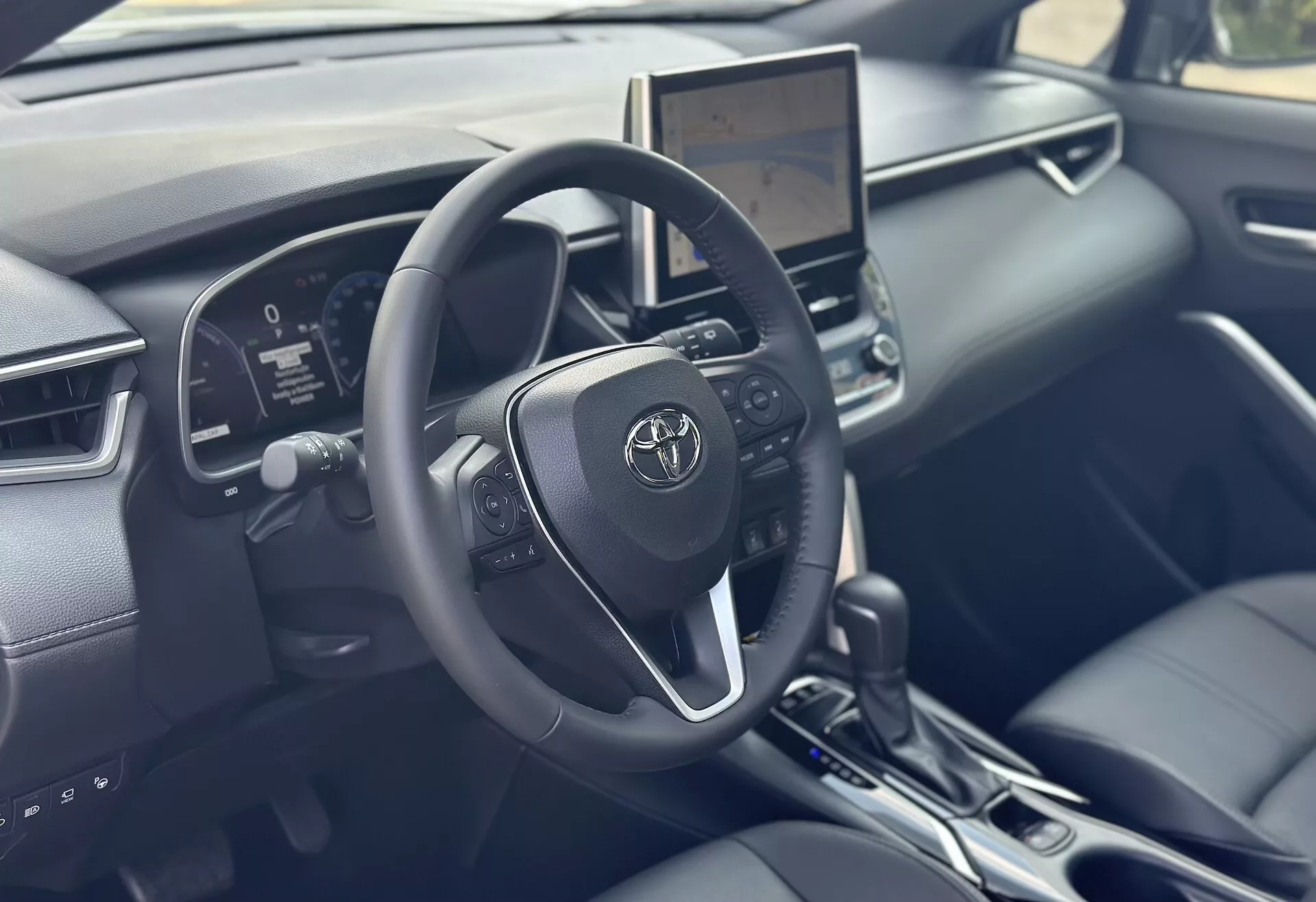 Interiér Toyoty Corolla Cross 2022 se zaměřením na stranu řidiče s detailním volantem, digitálním přístrojovým panelem a centrálním dotykovým infotainmentem s moderní technologií a akcenty z černé kůže