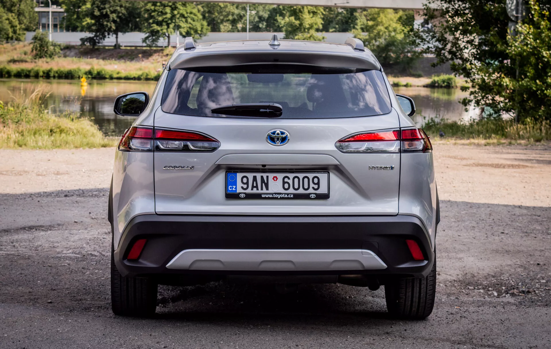 Zadní pohled na Toyota Corolla Cross 2022, zobrazující LED zadní světla, odznak hybrid a chromovaný emblém Toyota s přirozeným denním osvětlením a malebným pozadím zeleně a vody