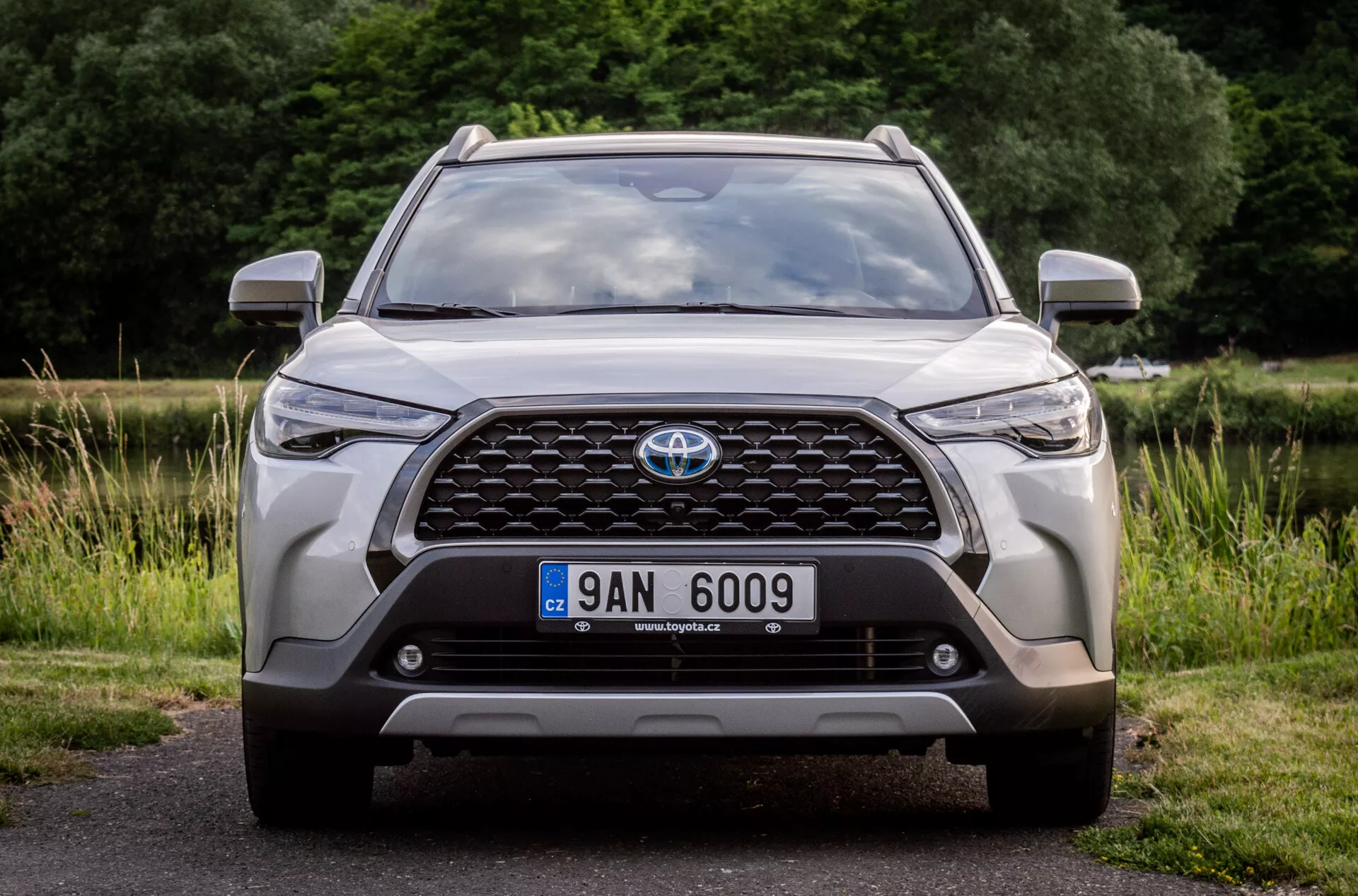 Přední pohled na Toyota Corolla Cross 2022, zdůrazňující jeho velkou mřížku s emblémem, moderní LED světlomety, mlhová světla, pozici registrační značky, integrované parkovací senzory nebo přední kameru, aerodynamické obrysy kapoty, robustní design spodní části nárazníku a sání vzduchu, zaparkovaný na asfaltu s přírodní kulisu