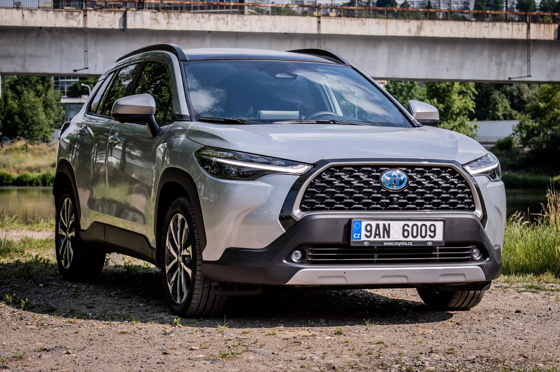 Toyota Corolla Cross 2022 zaparkovaná na štěrku ukazuje svůj robustní design, sportovní kola z lehké slitiny s více paprsky, pokročilé LED světlomety a vysokou světlou výšku z pohledu ze tří čtvrtě na přední mřížku, nárazník a prvky na straně řidiče s mostem na pozadí v poloměstském prostředí
