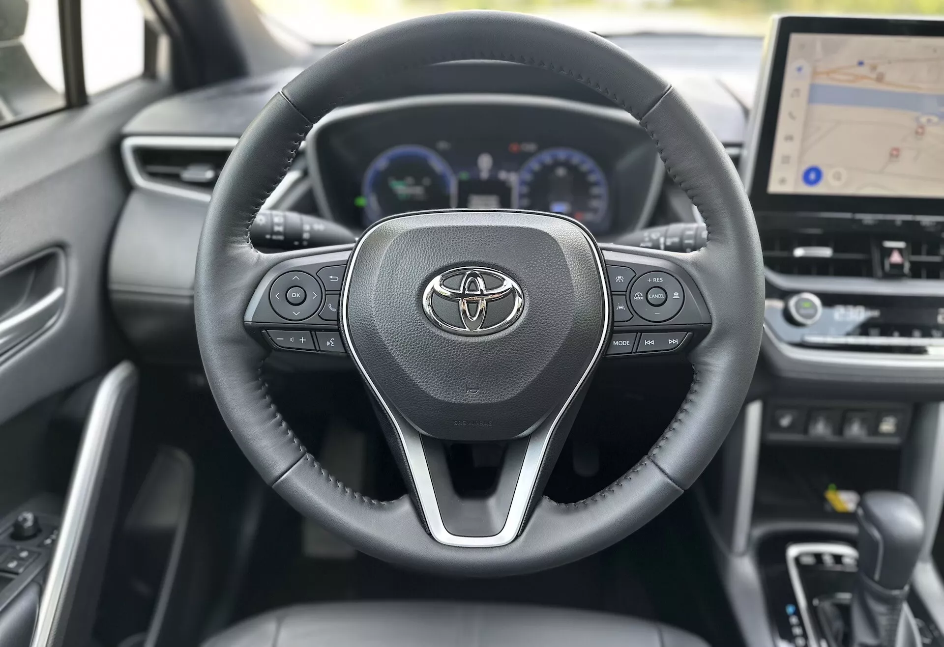 Interiér Toyoty Corolla Cross 2022 s volantem s logem Toyoty, integrovanými ovladači pro audio, hovory a tempomat, za kterým je částečně viditelný digitální palubní deska se speedometrem, otáčkoměrem a multifunkčním displejem, včetně většího centrálního displeje palubní desky pro GPS navigaci, vše na pozadí současného designu z tmavého materiálu.