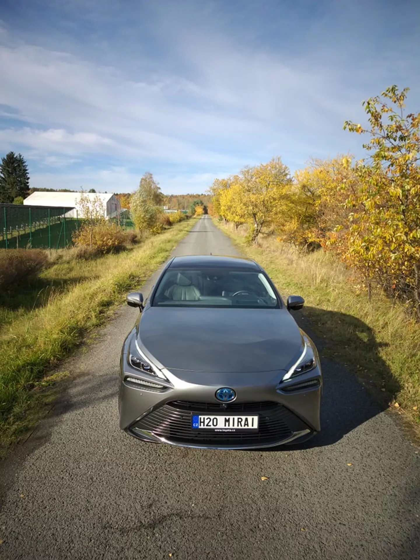 Toyota Mirai z roku 2021 fotografovaná z předního pohledu pod mírným úhlem, zobrazující její elegantní LED světlomety a výraznou masku s centrálním emblémem, který může zahrnovat technologii senzorů, na pozadí pololidké krajiny při přirozeném denním světle, předvádějící pokročilou technologii na vodíkové palivové články a aerodynamický design