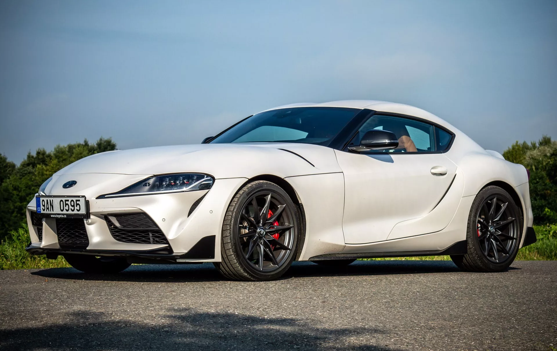 Bílé sportovní kupé Toyota Supra (GR-Supra) z roku 2019 zaparkované na asfaltu pod jasnou oblohou, boční profil zobrazující LED světlomety, sportovně aerodynamický design a výkonnostně orientované funkce