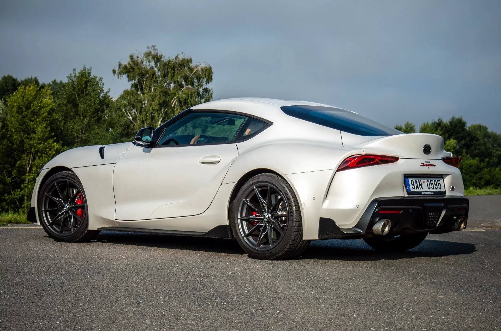 Toyota Supra (GR-Supra) z roku 2019 v bílé barvě předvádějící svůj aerodynamický design, sportovní postoj s zadním spoilerem, výkonné černé kola s červenými brzdovými třmeny, dvojitý výfukový systém a moderní LED zadní světla z bočně-zadní perspektivy