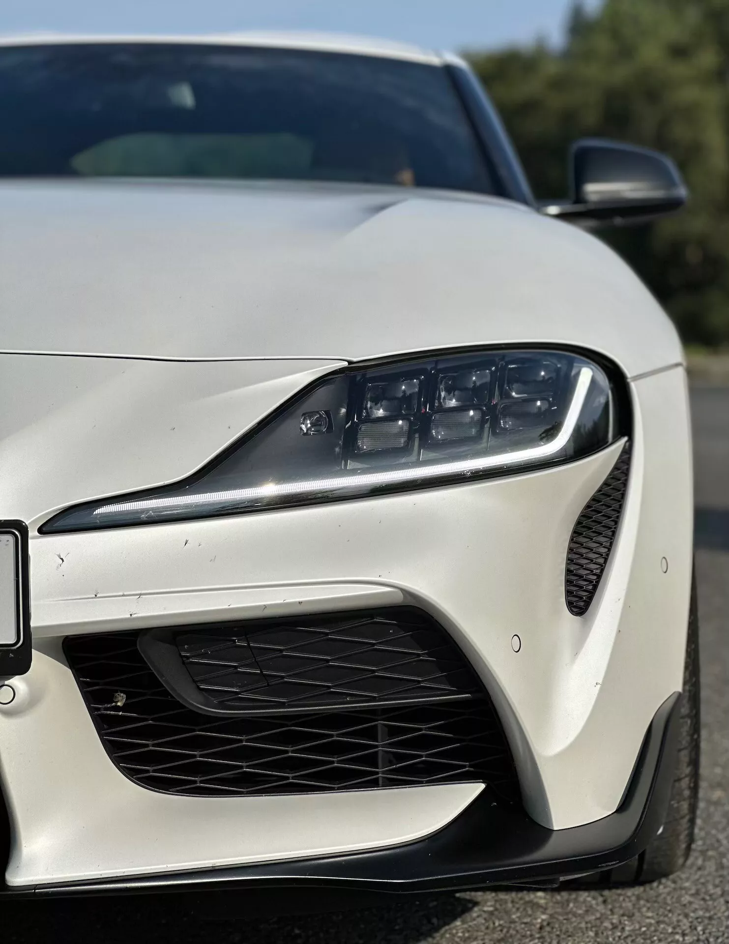 Detailní úhlový pohled zepředu vlevo na Toyota Supra (GR-Supra) z roku 2019 s moderním LED světlometem, výkonnostně orientovaným předním nárazníkem s mřížkou včelí plástve na sání vzduchu, aerodynamickými konturami a elegantním bočním zrcátkem na lesklém laku