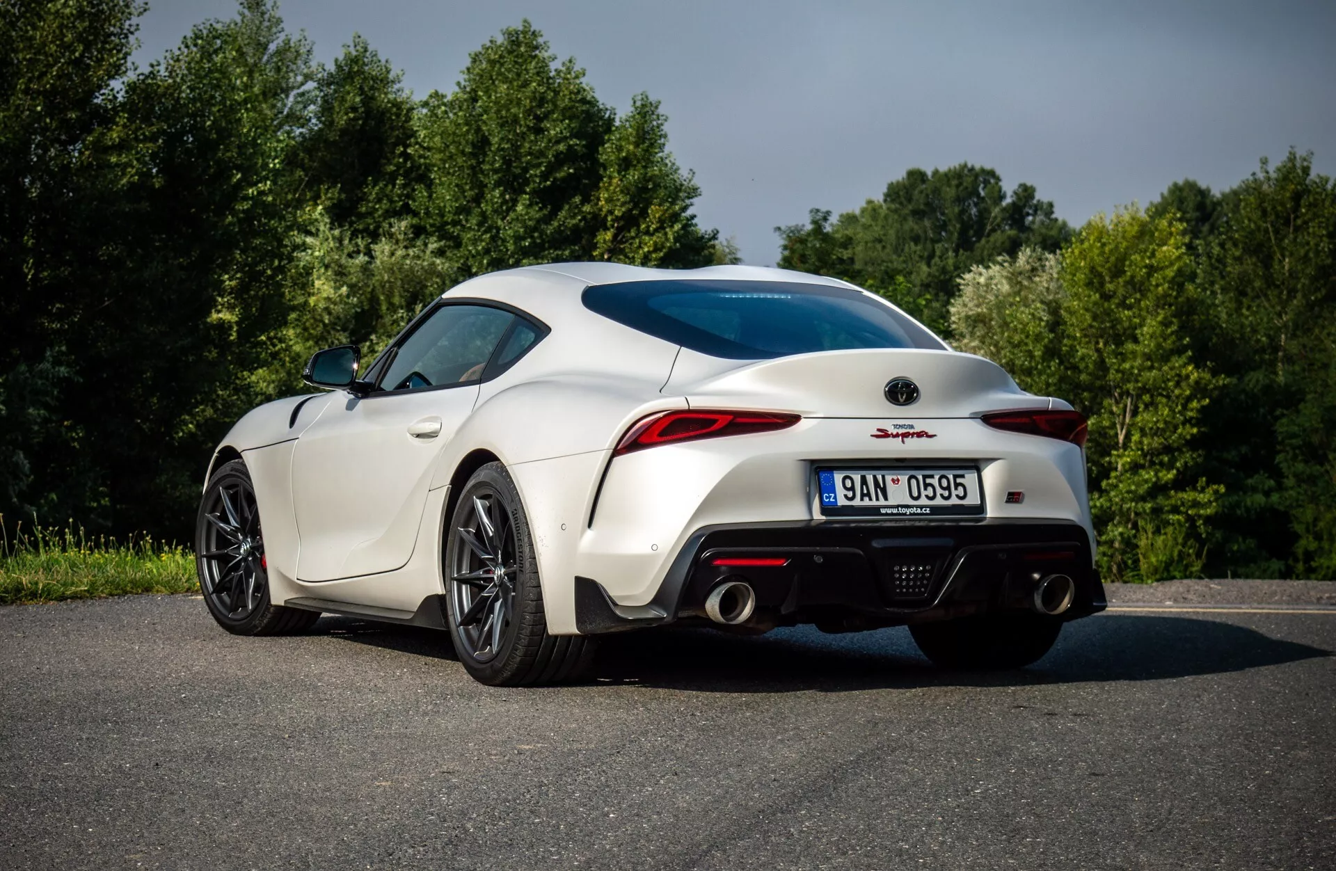 Bílý GR-Supra 2019, zobrazený z zadní trojčtvrteční perspektivy, ukazuje sportovní design s zakřivenými zadními částmi, originálními ráfky kol, bočními zrcátky, LED zadními světly, dvojitými výfuky a integrovaným spoilerem, obklopen zelení.