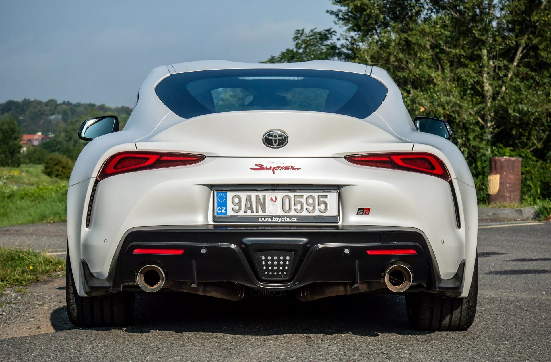 Zadní pohled na Toyota Supra (GR Supra) 2019 ukazující LED zadní světla, dvojitý výfukový systém, zadní difuzor, parkovací senzory a integrované křídlo na bílém reflexním povrchu, zaparkovaná na asfaltu s přírodní a venkovskou kulisou