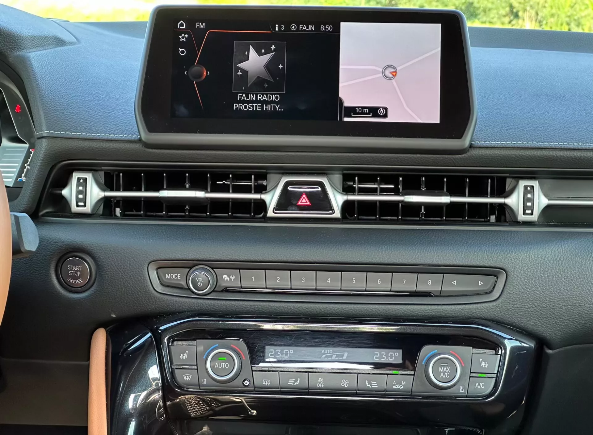 Vnitřní pohled na kokpit Toyota Supra GR 2019 zobrazující centrální konzolu s digitálním dotykovým infotainment displejem s 'FAN RADIO' a navigací mapy, řadou vzduchových ventilů, tlačítkem výstražných světel, tlačítky médií a klimatizace s nastavením dvojzónové teploty na 23°C a elegantními designovými prvky kontrastního obložení.
