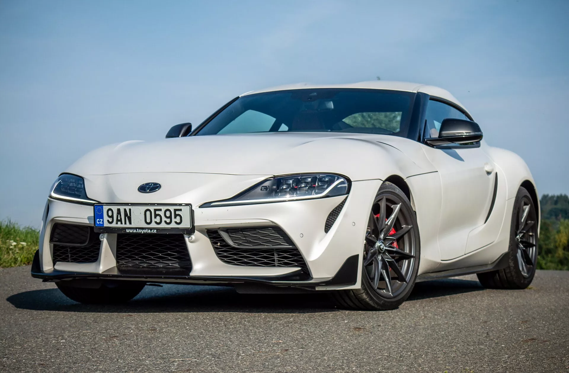 Bílý Toyota Supra GR 2019 zobrazený z nízkého předního tříčtvrtečního úhlu, zdůrazňující jeho aerodynamický profil, úzké LED světlomety, velké vzduchové vstupy, přední splitter a sportovně tvarovanou kapotu.