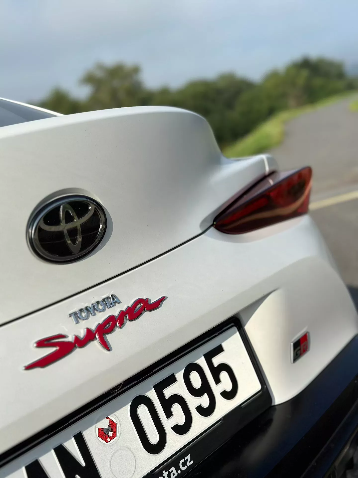 Detailní záběr zadní části bílé Toyoty Supra 2019 s logem Toyota, červeným nápisem modelu 'Supra', pravým LED zadním světlem, zapuštěnou oblastí pro poznávací značku, částí zadního nárazníku, integrovaným spoilerem, částí střechy a zadním čelním sklem na pozadí rozmazané silnice a zeleně.