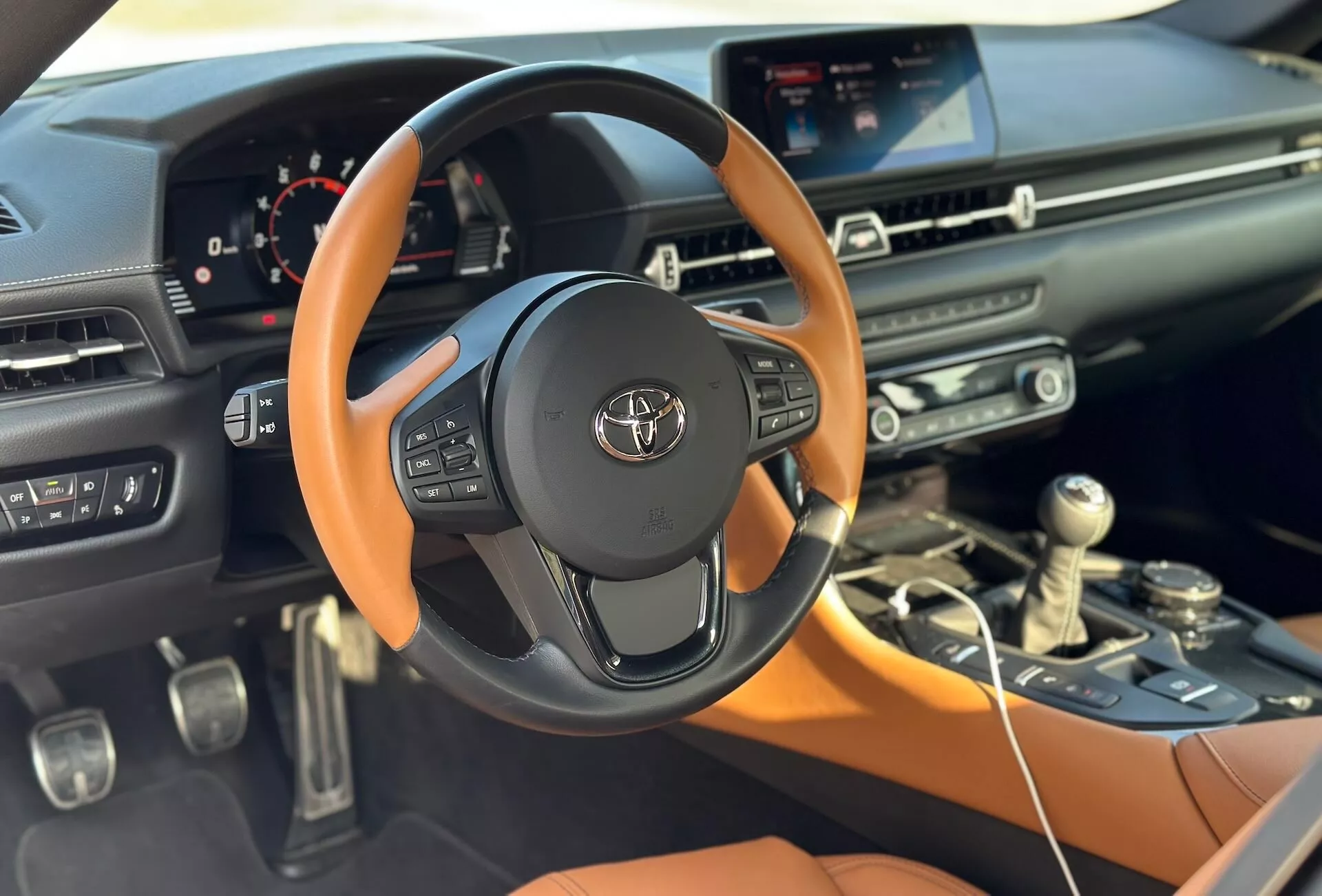 Detailní pohled na interiér Toyoty Supra 2019 GR-Supra ukazující volant s emblémem Toyota, středovou konzolu, dotykový displej infotainmentu a částečný palubní desku spolujezdce se zaměřením na technologické a designové prvky