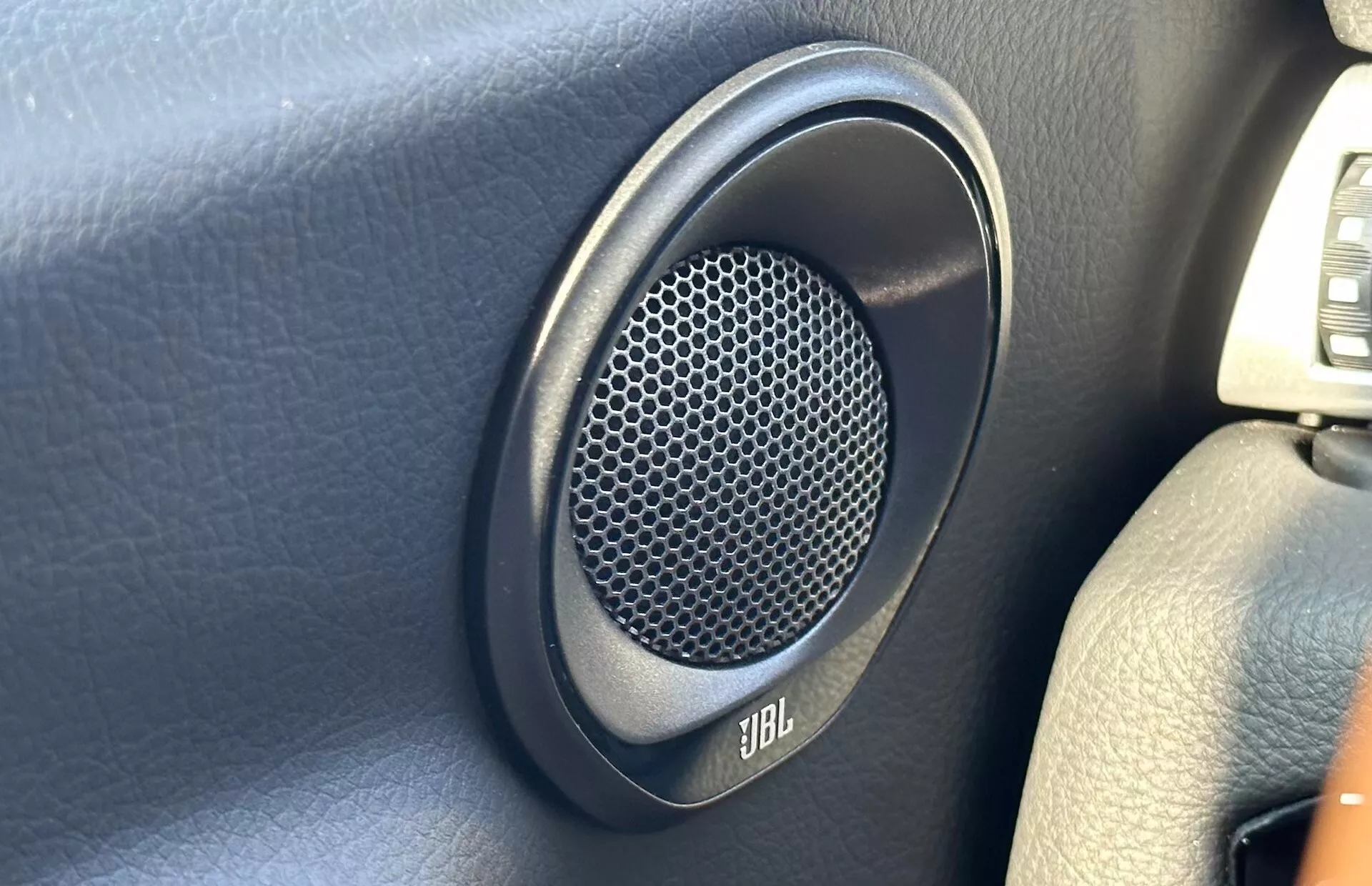 Detailní pohled na reproduktor značky JBL umístěný na tmavém panelu dveří Toyoty Supra 2019, s šestiúhelníkovou včelí pláství mřížkou pro lepší zvukový výkon.