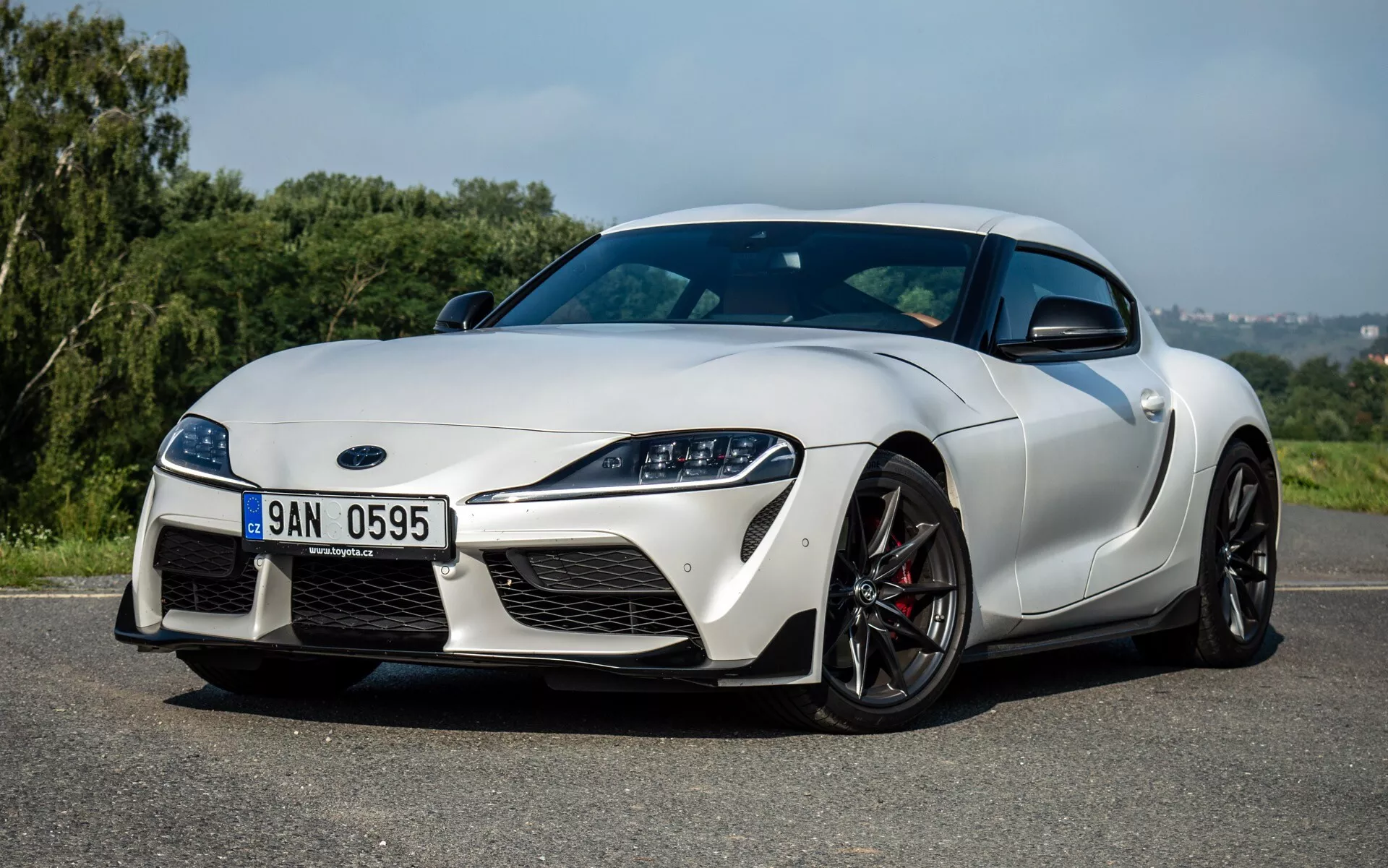 Sportovní auto Toyota GR Supra 2019 v pohledu z předního tříčtvrtečního úhlu ukazující aerodynamický design, elegantní přední mřížku, černá kola s vícepaprskovým designem a lesklý bílý lak na asfaltu s pozadím zeleně
