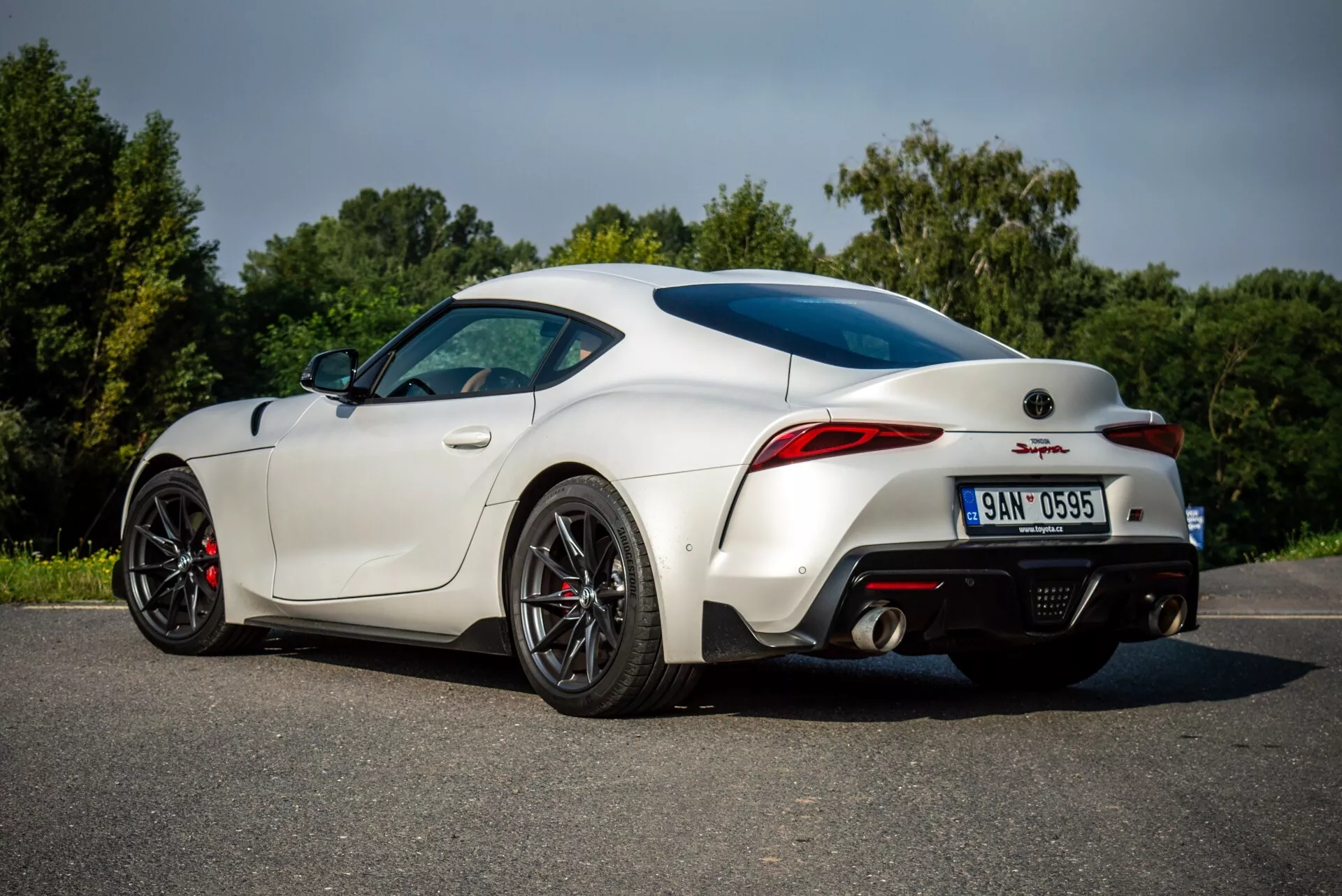 Bílý Toyota Supra GR 2019 ukazuje svůj aerodynamický design z pohledu ze zadu a strany pasažéra na asfaltu, včetně charakteristických LED zadních světel, výkonnostních kol s červenými brzdovými třmeny, moderního výfukového systému a zadního difuzoru pro lepší aerodynamiku.