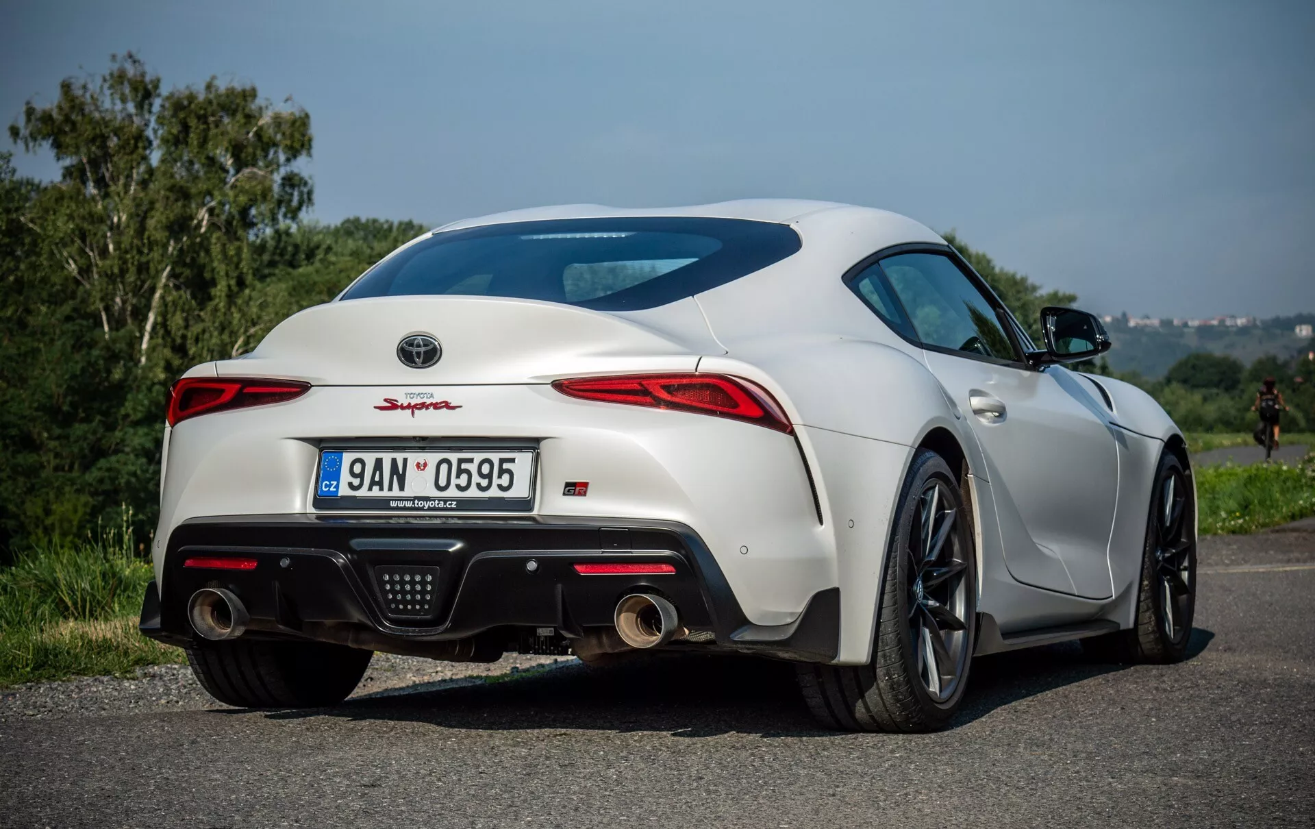 Toyota Supra 2019 v jasně bílé barvě, předvádí sportovní a aerodynamický design kupé z pohledu ze zadní tříčtvrteční perspektivy, s černými slitinovými koly s více paprsky a výkonnými pneumatikami, charakteristickými LED zadními světly, integrovaným zadním spoilerem pro aerodynamiku a dvojitým výfukovým systémem, zaparkovaná venku s čistou oblohou na pozadí.
