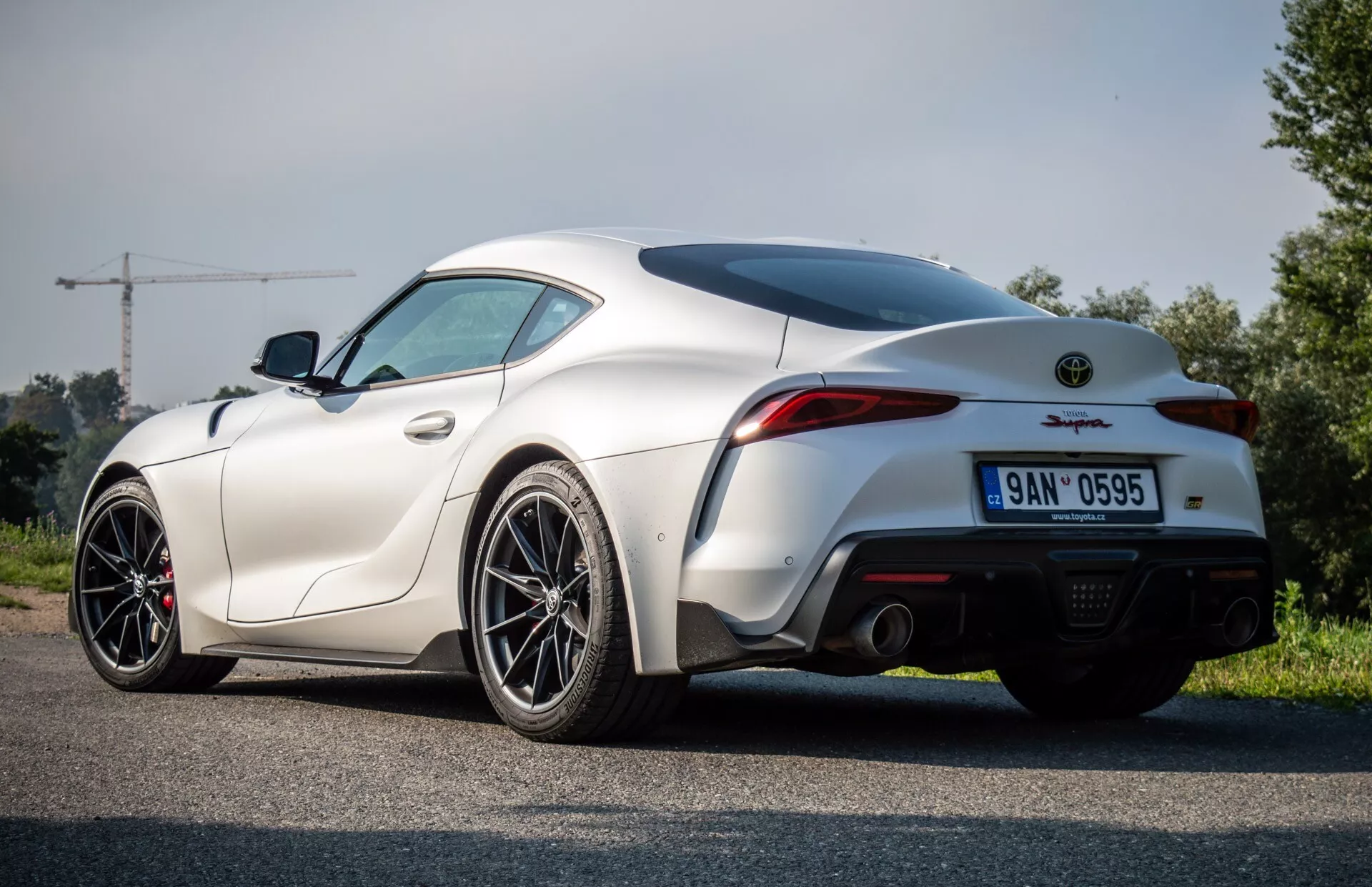 Toyota Supra GR z roku 2019, bílá, zobrazení ze zadního tříčtvrtečního úhlu na asfaltové cestě, zdůrazňující křivku zadního blatníku, design kufru a nárazníku s dvojitým výfukem, ve venkovském prostředí
