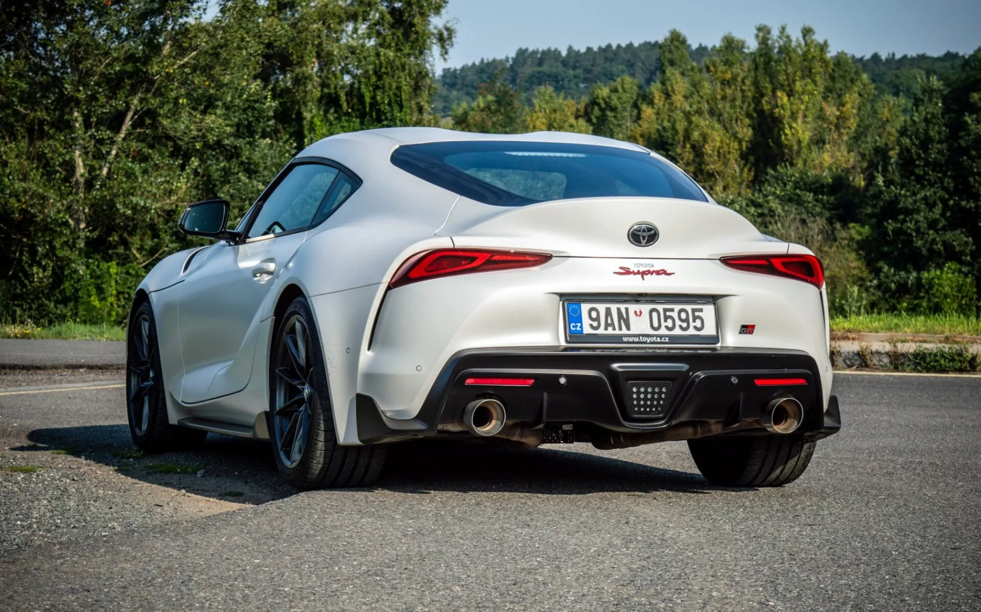 Bílý Toyota Supra (GR Supra) z roku 2019 se zobrazením zezadu na tři čtvrtiny s důrazem na aerodynamický design včetně zadního spoileru, LED zadních světel a sportovního zadního difuzoru na asfaltovém povrchu
