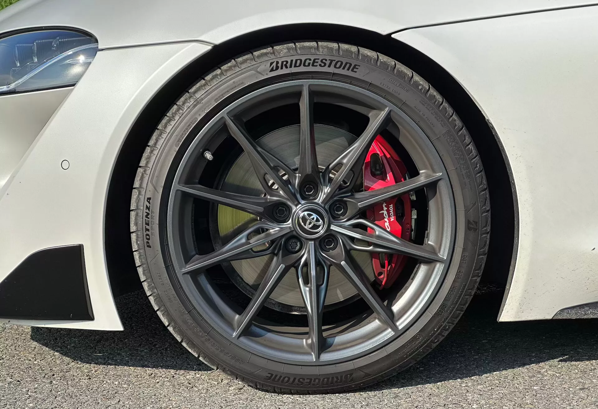 Detail předního kola na straně řidiče Toyota Supra (GR Supra) z roku 2019, zobrazující 10ramenný tmavě kovový slitinový ráfek s výkonnostní pneumatikou Bridgestone POTENZA, 255/35 R19, včetně červeného výkonnostního brzdového třmenu a ventilovaného disku, částečný pohled na karoserii s kovovým lakováním