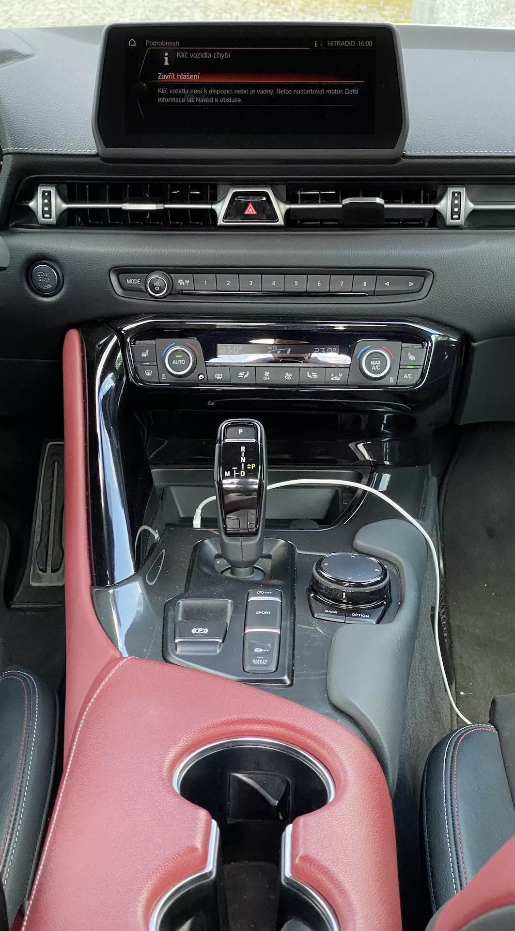 Svislý snímek z pohledu řidiče nebo spolujezdce, který zvýrazňuje středovou konzoli a palubní desku vozu Toyota Supra GR 2.0 Pure Automatic z roku 2019. V horní části palubní desky je displej infotainmentu v režimu standby, pod kterým jsou hmatová tlačítka a střední výdech vzduchu. Níže jsou tlačítka a knoflíky pro ovládání klimatizace s kovovým povrchem. Středová konzole je částečně pokryta materiálem podobným červené kůži s lesklým černým lemováním, včetně řadicí páky s koženým potahem pro automatickou převodovku a tlačítek pro jízdní režimy, držáku na nápoje a malé úložné oblasti. Opěrka, lehce rozostřená, je také pokryta červeným materiálem. Celkový interiér naznačuje kombinaci moderní technologie a tradičních fyzických ovladačů s prémiovými sportovními úpravy.