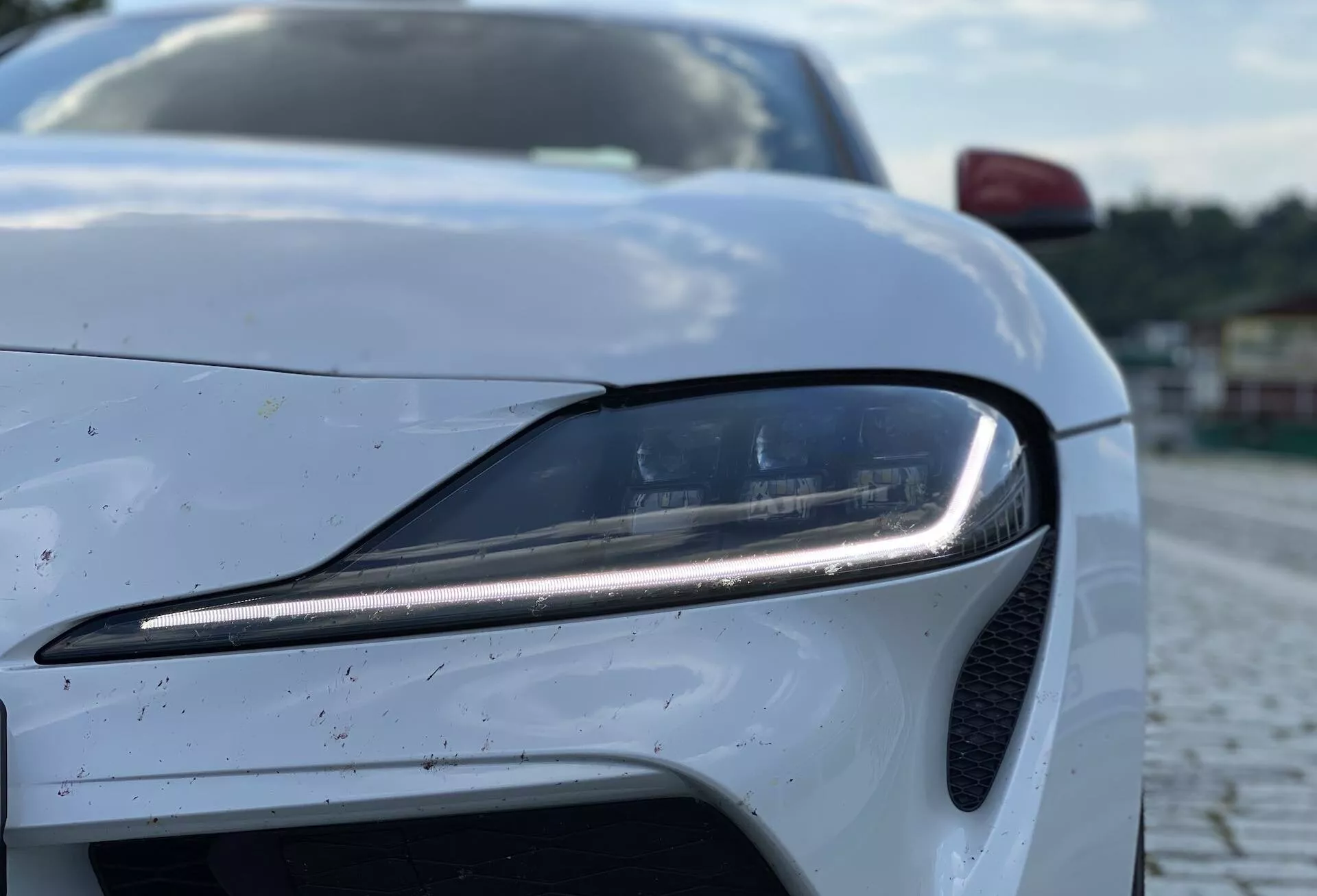 Detailní pohled na Toyota GR Supra 2.0 Pure Automatic z roku 2019, zaměřený na moderní LED přední světlo na straně řidiče a část mřížky. Fotografie je pořízena z nízkého úhlu lehce nahoru, čímž se zdůrazňuje solidní přítomnost a sportovní design auta. Bílý lak je posetý malými špínami nebo kapkami deště, což naznačuje nedávné použití. Kompozice se soustředí na světlomet s rozostřeným pozadím, zvýrazňující pokročilý osvětlovací systém a aerodynamické prvky.