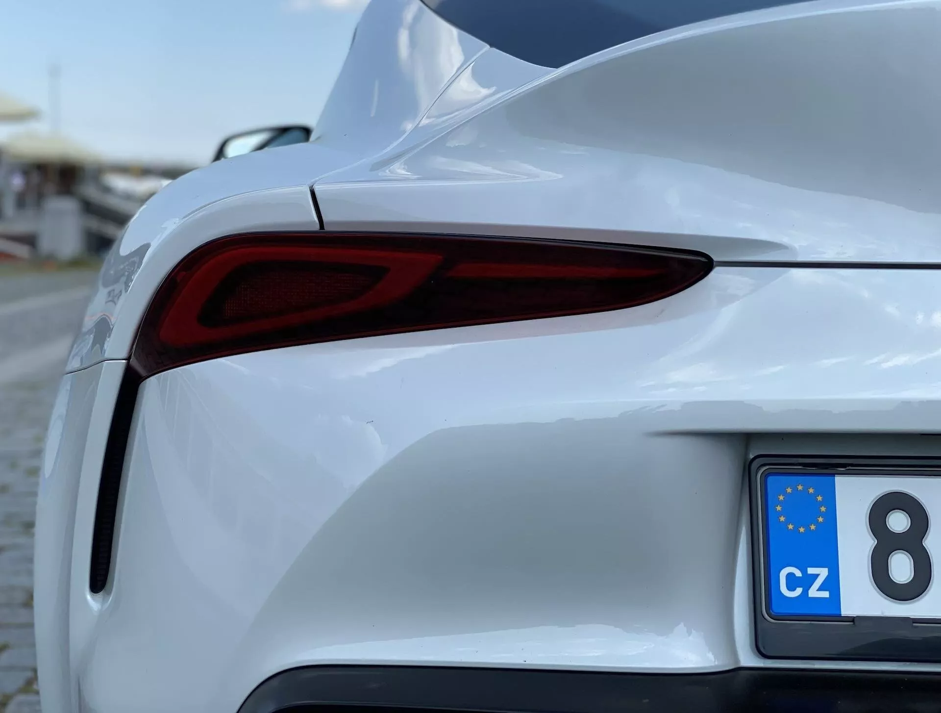 Detailní částečný pohled na bílý Toyota Supra 2019 (toyota-gr-supra-2-0-pure-automatic), zaměřený na zadní pravou část z nízkého, necentrovaného úhlu. Obrázek ukazuje elegantní LED zadní světla, sportovně laděný zadní nárazník, lesklý lak a registrační značku s EU znakem z České republiky.