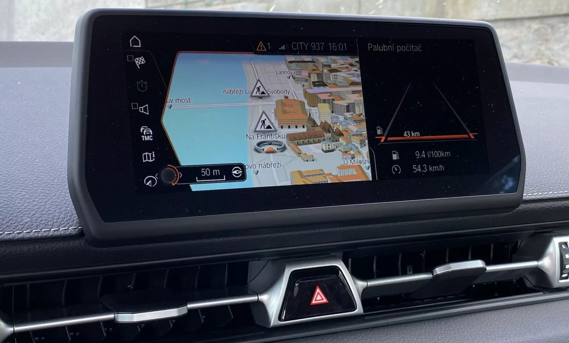 Obrázek představuje centrální konzolu Toyoty GR Supra 2.0 Pure Automatic z roku 2019, zaměřující se na vestavěný infotainment systém s výrazným navigačním displejem. Fotografováno mírně z pravého úhlu, zobrazuje navigační obrazovku s 3D mapou ulice, budov a různých ikon pro navigační pokyny a body zájmu. Zobrazena je trasa a indikátor polohy vozidla s ukazateli vzdálenosti a směrovými pokyny. Pod obrazovkou je řada z větší části zakrytých tlačítek se známým červeným trojúhelníkem ochranného varování. Po stranách obrazovky jsou mřížky nebo otvory, naznačující integraci s klimatizačním systémem. Detailní záběr zdůrazňuje pokročilé automobilové technologie a funkce infotainmentu, jako jsou audio zábava, možnosti připojení a nastavení vozidla, bez zobrazení exteriéru nebo zbytku interiéru.