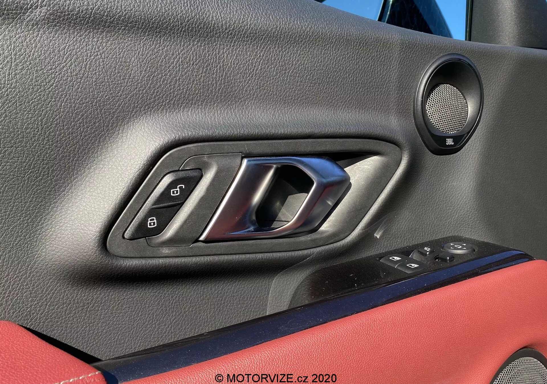 Detailní pohled na vnitřek dveří řidiče vozu Toyota GR Supra 2019 (2.0 Pure Automatic), obsahující kovovou klikou umístěnou v černém plastu, ovládací panel s elektrickými okny a zrcátky, elektronickým zámkem, viditelnou mřížku reproduktoru a kombinaci textur včetně červeného akcentu na panelu dveří. Důraz na moderní design a funkčnost interiéru vozu.