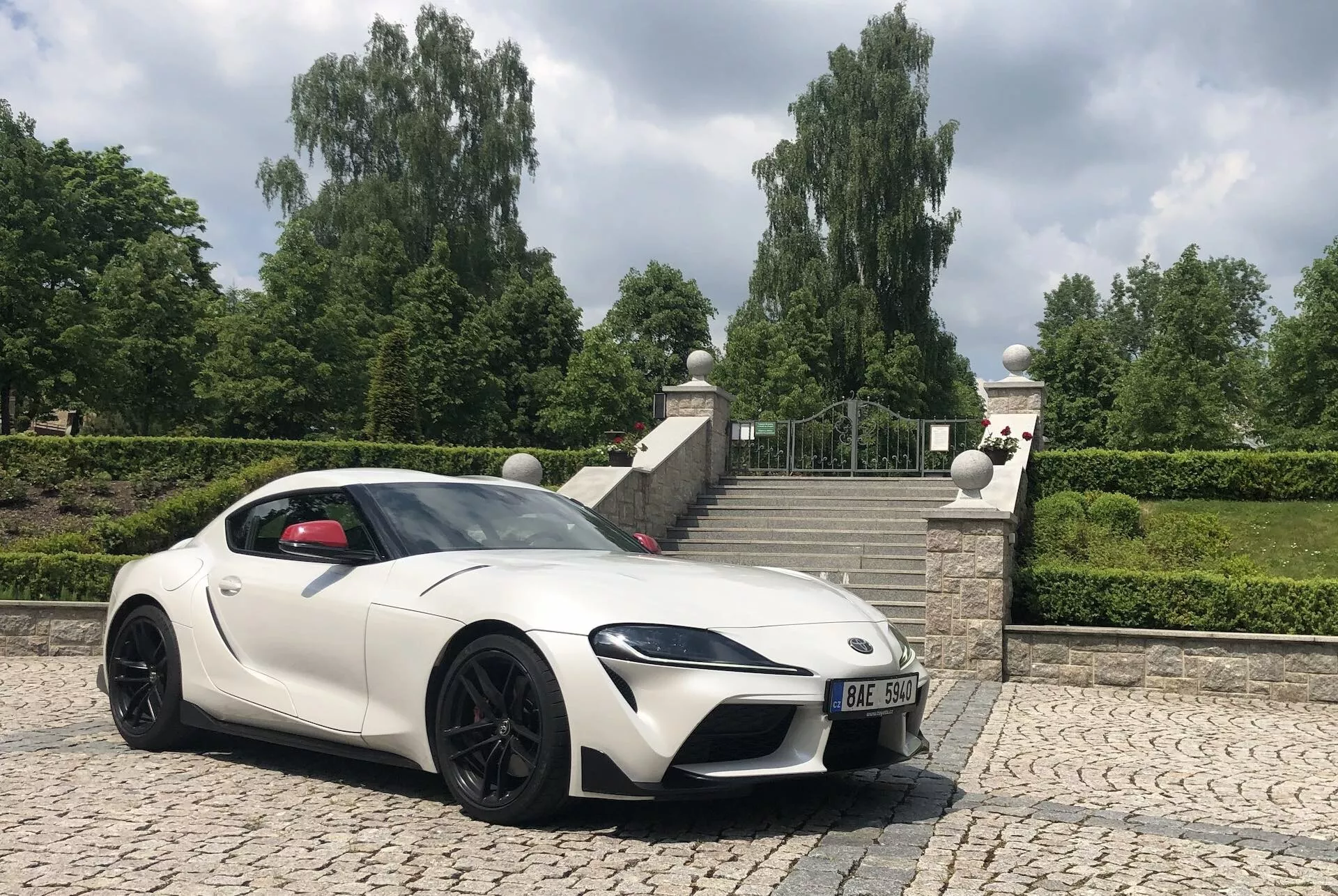 Přední tříčtvrtinový pohled na bílý sportovní automobil Toyota GR Supra 2.0 Pure Automatic z roku 2019 na dlážděném povrchu, fotografovaný z nízkého úhlu s jasnou oblohou a vegetací v pozadí. Obrázek zvýrazňuje agresivní postoj automobilu, sportovní linie, ostře navržené LED světlomety, velkou přední mřížku pro aerodynamiku a chlazení motoru, estetické a funkční větrací otvory na kapotě, elegantní aerodynamický boční profil, designová boční zrcátka, velká slitá kola s výkonnými brzdami a nízkoprofilové pneumatiky. Dlouhá kapota a kabina posunutá směrem dozadu zdůrazňují uspořádání s předním motorem a pohonem zadních kol.