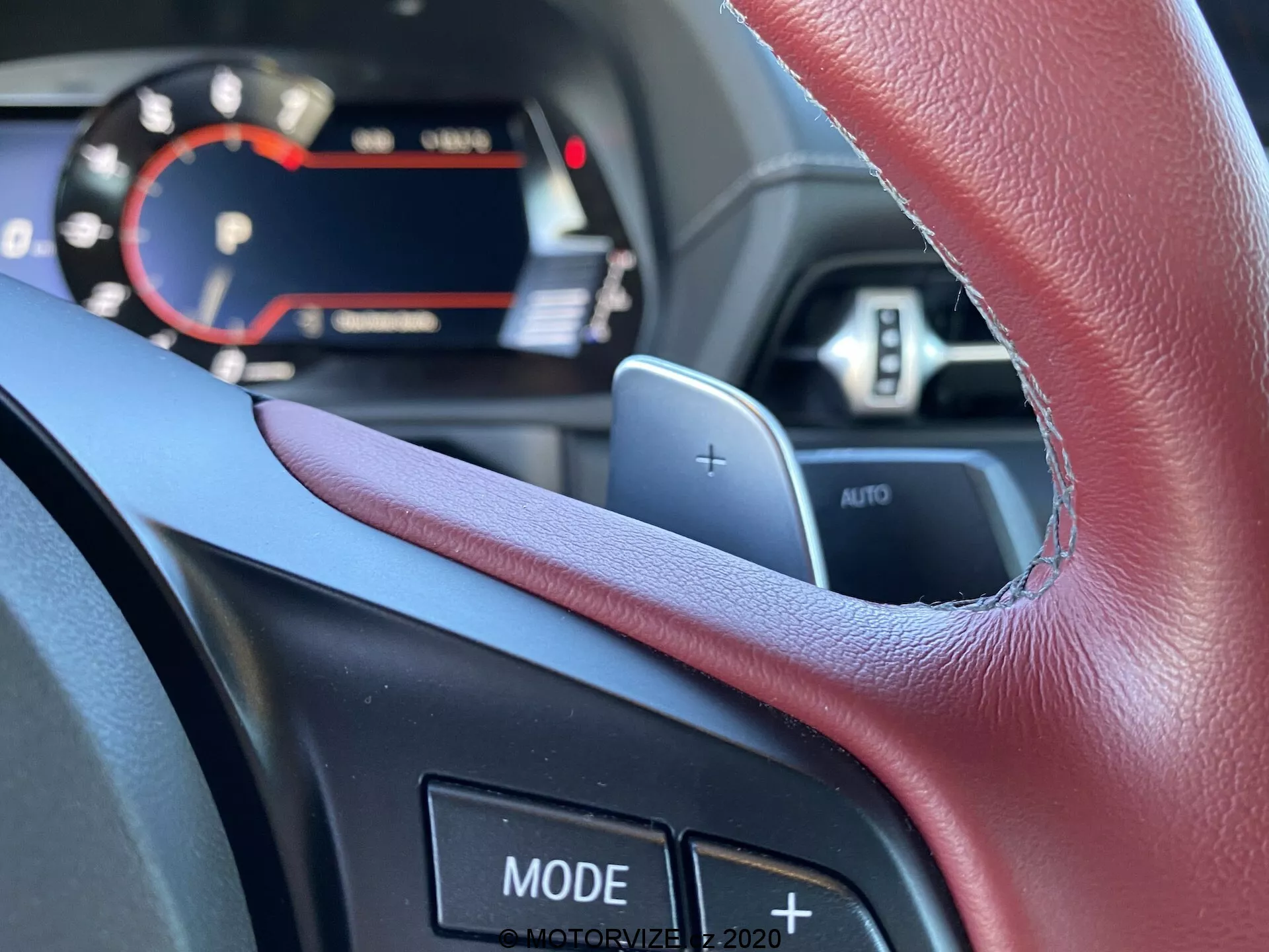 Pohled z pohledu řidiče na interiér Toyoty Supra 2019, zaměřující se na multifunkční volant s páčkovým řazením a různými ovládacími tlačítky. Část digitálního panelu přístrojů s moderními displejovými prvky je viditelná přes horní okraj volantu a zdůrazňuje pokročilé technologické vlastnosti vozu.