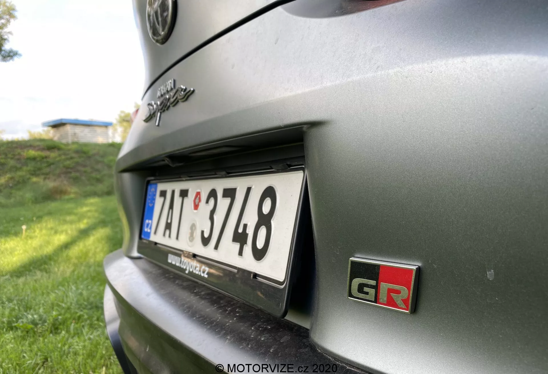 Blízký pohled na zadní část kovově šedého vozu Toyota GR Supra 2.0 Pure Automatic z roku 2019. Zaměřuje se na oblast poznávací značky a okolní detaily, se zdůrazněním pravé strany zadní části vozu. Pohled je z nízkého úhlu, jakoby z pozice přikrčené osoby, mírně vzhůru směrem k zadní části vozidla. Výrazným prvkem je odznak 'GR', což značí výkonově orientovanou úpravu od Toyota's Gazoo Racing, a evropský styl poznávací značky. Hloubka ostrosti je mělká, s ostrým zaměřením na poznávací značku a znak GR, zatímco zbytek karoserie vozu a pozadí jsou rozmazané. Osvětlení naznačuje, že snímek byl pořízen venku za jasného dne, s přirozeným světlem vytvářejícím stíny a zvýraznění na povrchu vozu.