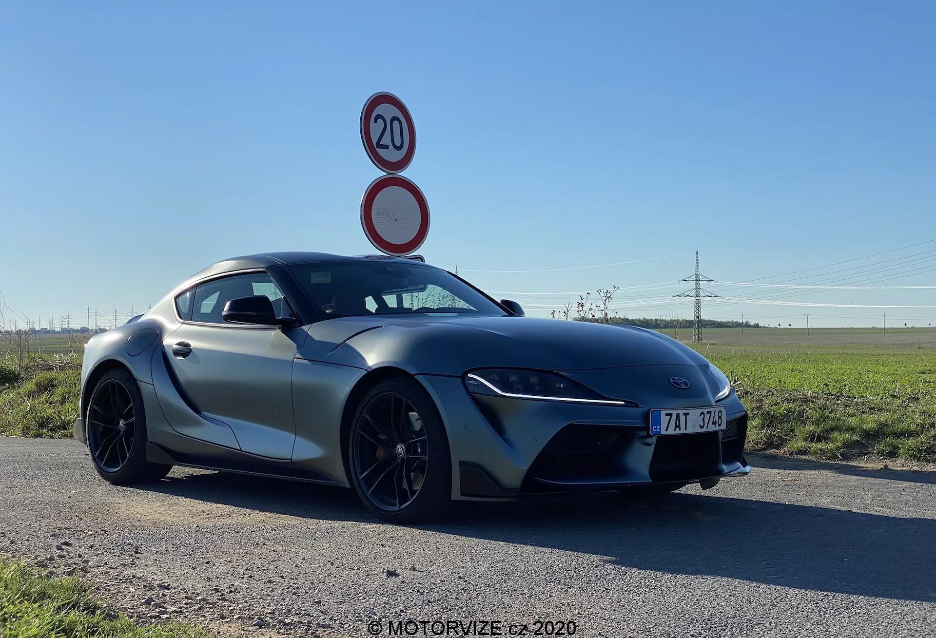 Obrázek ukazuje sportovní vůz Toyota GR Supra 2.0 Pure Automatic z roku 2019 z nízkého tříčtvrtečního úhlu zepředu, zdůrazňuje jeho elegantní, aerodynamické linie, úzké LED světlomety a velký přední nasávací otvor. Kapota se svažuje do sportovní přední masky, naznačující přední uspořádání motoru. Boční zrcátka jsou namontována na dveřích, zvýrazňující jeho střechu ve stylu fastback, která zvýrazňuje vzhled kupé. Pozoruhodné rysy zahrnují vícepaprsková černá kola s pneumatikami s nízkým profilem a ventilovaný brzdový systém, který naznačuje vysoký výkon. Vůz je umístěn na asfaltové silnici proti plochému venkovskému pozadí, což zdůrazňuje jeho pokročilý design.
