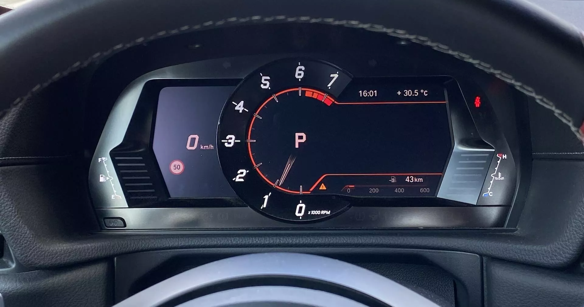 Pohled řidiče na digitální přístrojovou desku vozu Toyota GR Supra 2.0 Pure Automatic 2019. Centrální otáčkoměr s červenou ručičkou na '0' ot./min ukazuje nečinný motor; červená kontrolka 'P' svítí pro parkovací režim. Levý displej zobrazuje ikonu vozidla a možné výstražné kontrolky pro otevřené dveře. Rychloměr není jasně vidět, pravděpodobně na digitálním displeji. Vpravo palivoměr a teploměr motoru, s palivem nad čtvrtinou a teplotou v normálním rozsahu. Další detaily zahrnují venkovní teplotu (30,5°C), čas (16:01) a stav tachometru (833 km), což naznačuje nízký počet najetých kilometrů. Viditelný vrch volantu naznačuje luxusní výbavu s detaily prošití. Pohled z první osoby zvýrazňuje moderní, pokročilou digitální zobrazovací technologii pro řidiče.