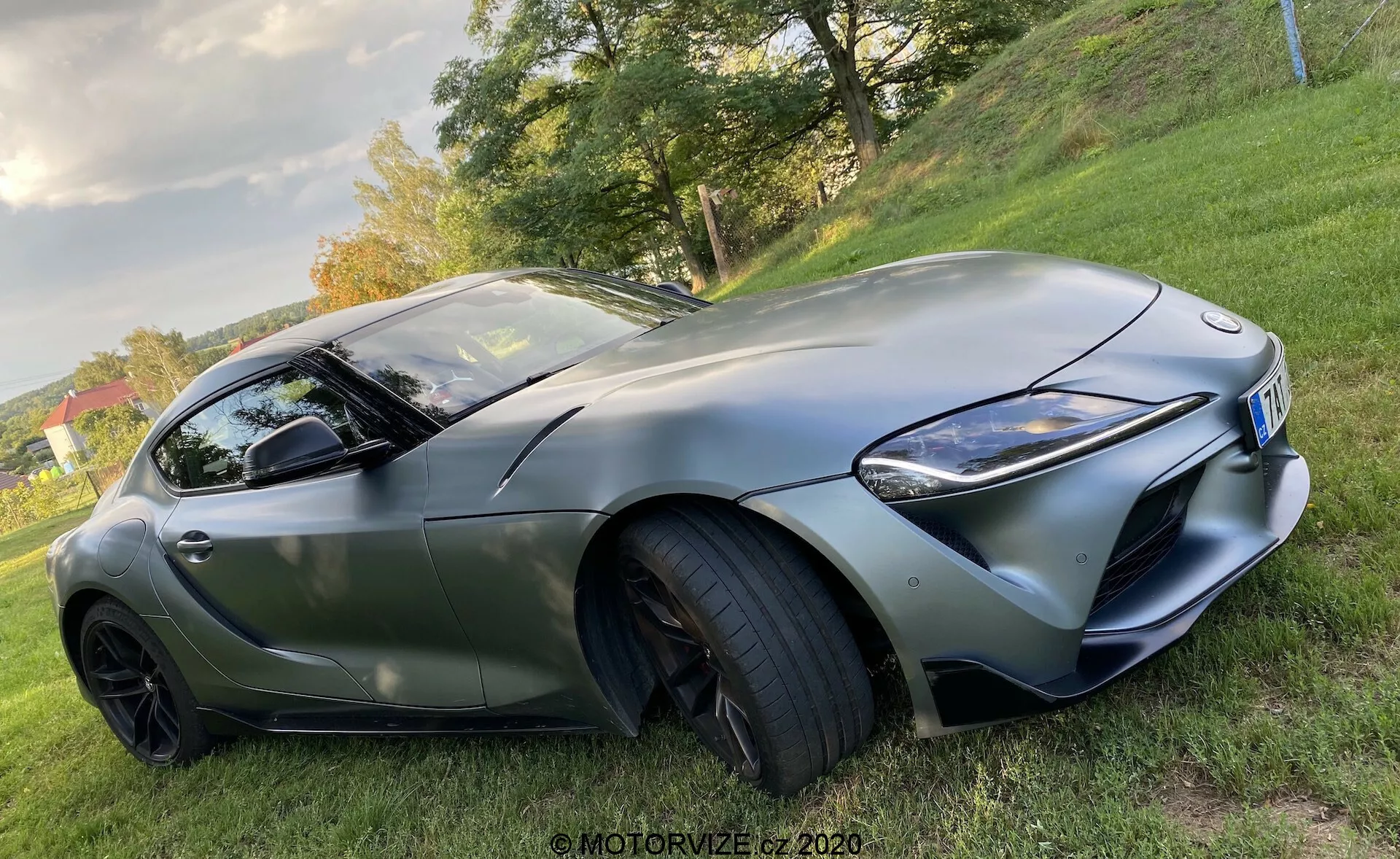 Pohled z nízkého úhlu na Toyota GR Supra 2.0 Pure Automatic z roku 2019, který ukazuje jeho sportovní siluetu z přední strany, zdůrazňující stranu řidiče. Snímek zvýrazňuje aerodynamické kontury vozu, výraznou přední masku, ostrá LED světla, velké nasávací otvory na předním nárazníku, tvarovanou kapotu, nízkoprofilové vysokovýkonné pneumatiky na víceramenových černých litých kolech, integrované směrovky v zrcátku a tónovaná okna s elegantní linií střechy a matným povrchem.