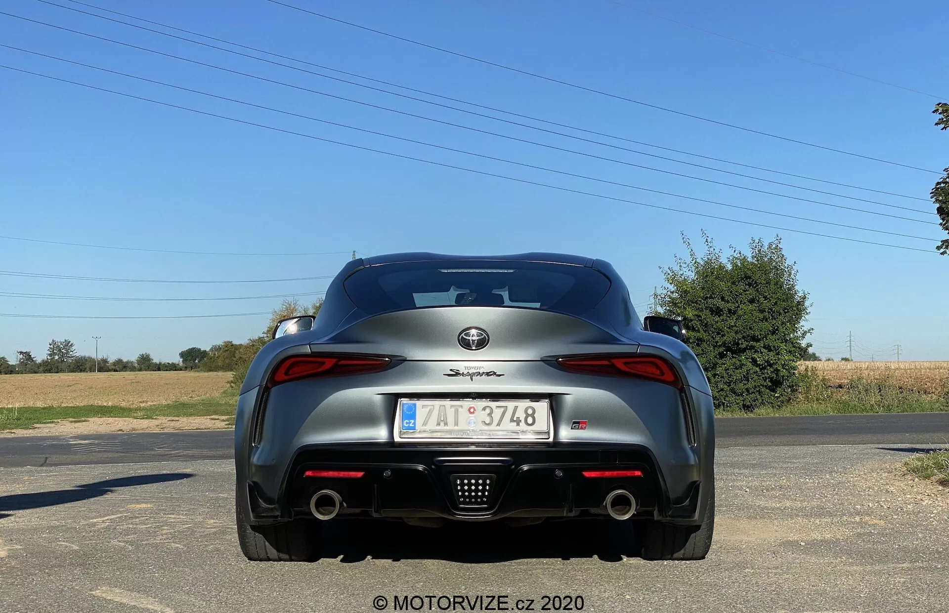 Pohled zezadu na Toyota Supra 2019 (Toyota GR Supra 2.0 Pure Automatic) jasného dne s venkovským pozadím. Fotografie z úrovně země ukazuje zadní část sportovního vozu v klidu. Viditelné designové prvky zahrnují výrobní odznak na víku kufru, modelový název nad poznávací značkou, výrazné zadní světlomety, dvojité výfukové trubky, aerodynamické linie s integrovaným zadním spojlerem a centrálně umístěnou poznávací značku s EU symbolem. Zadní sklo je mírně zakřivené a vůz je dokončen v tmavé, třpytivé barvě.