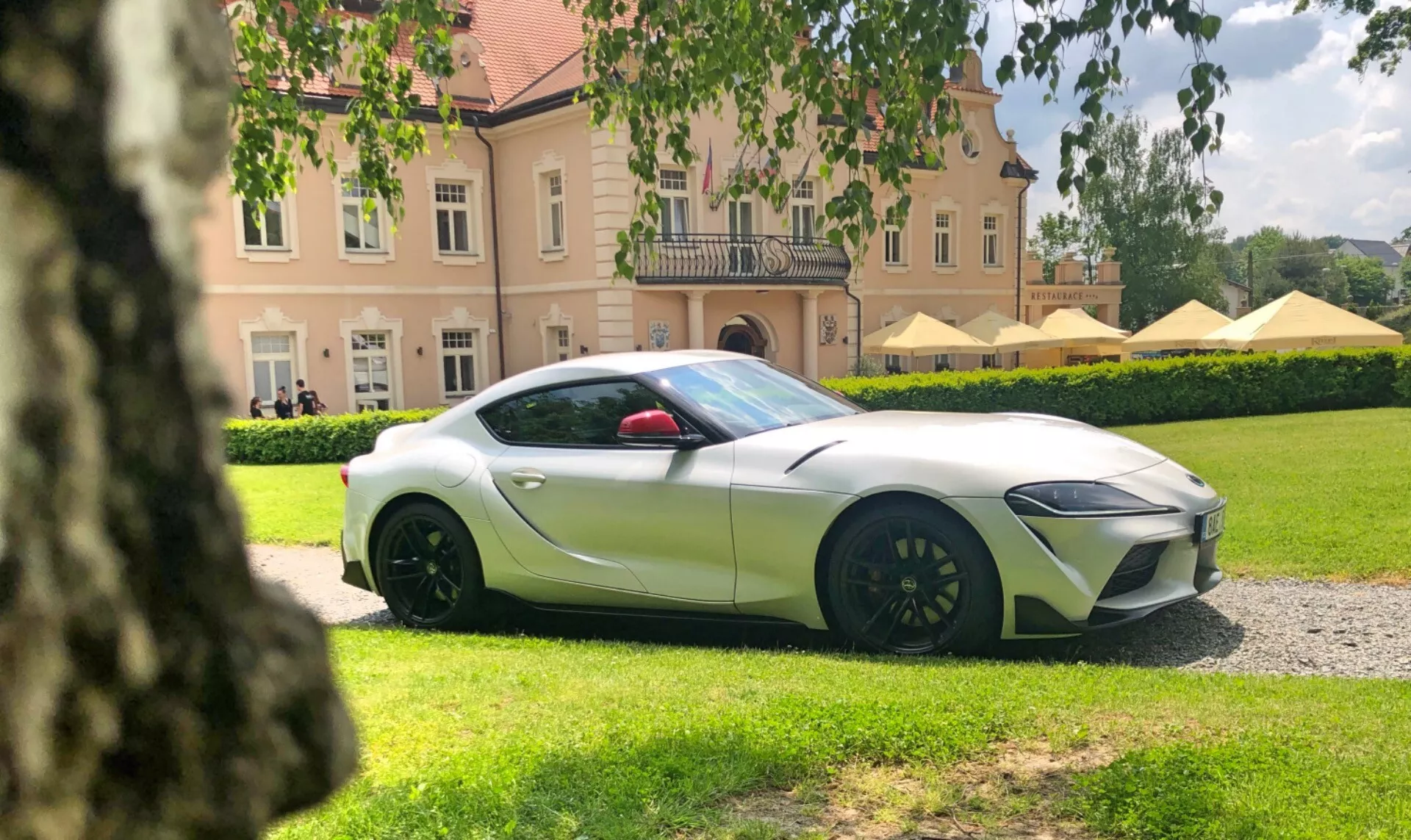 Obrázek Toyota Supra 2019 (Toyota-GR-Supra-2-0-Pure-Automatic), focený z nízkého bočního úhlu a zaměřený na stranu řidiče. Auto, natřené v metalické bílé s kontrastními kryty zrcátek, je umístěné přibližně 15 až 20 stop od kamery na štěrkové cestě, s pečlivě udržovaným trávníkem a velkou budovou v pozadí. Viditelný je přední levý kvadrant vozidla, ukazující ostré a úhlové světlomety, vícespětnicový design kol v tmavé barvě a značně velké brzdové třmeny, což naznačuje pokročilé automovilní technologie a vysoce výkonný brzdový systém.