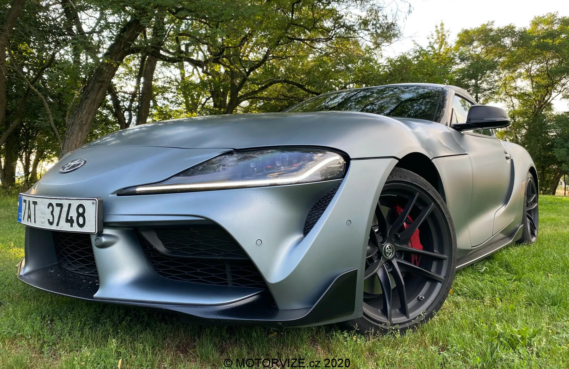 Sportovní vůz Toyota Supra 2019 zobrazený z nízkého, předního tříčtvrtečního úhlu, zaparkovaný na trávníku s pozadím stromů a listí. Dynamický a agresivní postoj zdůrazňuje jeho designové linie a aerodynamiku. Mezi vlastnosti patří výrazná přední maska, elegantní úhlová LED světla s denním svícením, dlouhá kapota s výraznými konturami, černá kola s mnoha paprsky s kontrastními červenými brzdovými třmeny, nízkoprofilové sportovní pneumatiky, nasávací otvor na předním blatníku a elegantní boční zrcátka.