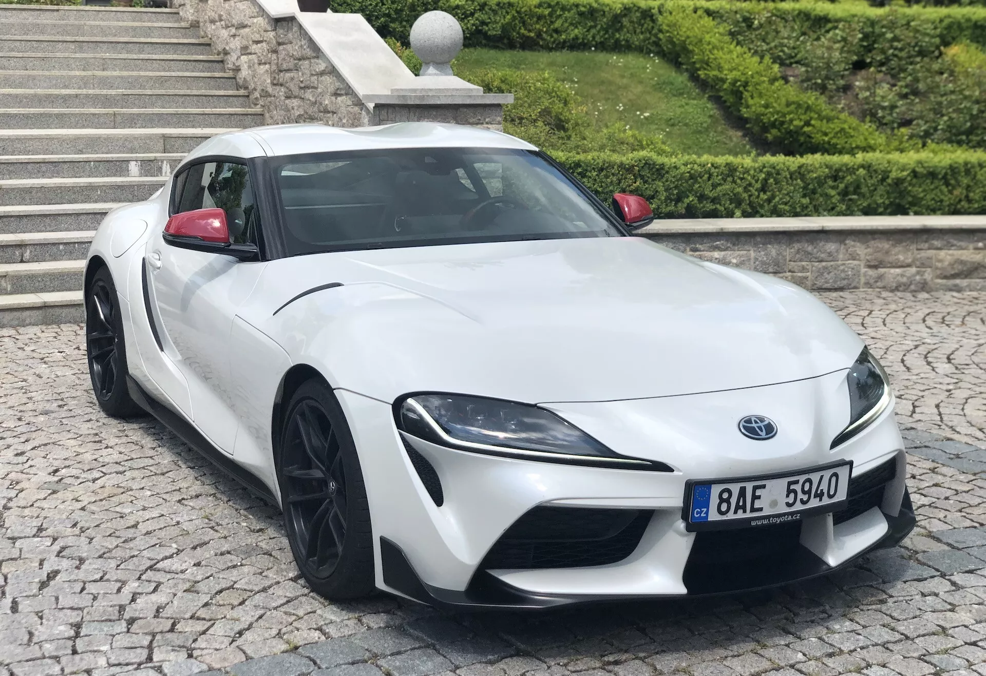 Obrázek zobrazuje moderní sportovní vůz Toyota Supra z roku 2019, zachycený z tříčtvrtečního pohledu zepředu. Auto má výraznou mřížku chladiče, tvarovaný nárazník s integrovanými otvory pro nasávání vzduchu, úzké LED světlomety s denními světly a konturovanou kapotu pro lepší aerodynamiku. Vozidlo je osazeno velkými černými víceramennými slitinovými koly, má hladké aerodynamické boční profily, kontrastní černou střechu proti bílé karosérii a tónovaná okna. Fotografie zaostřuje ostře na vozidlo s lehce rozostřeným pozadím, pořízená ve výšce kapoty, což činí vůz impozantním.