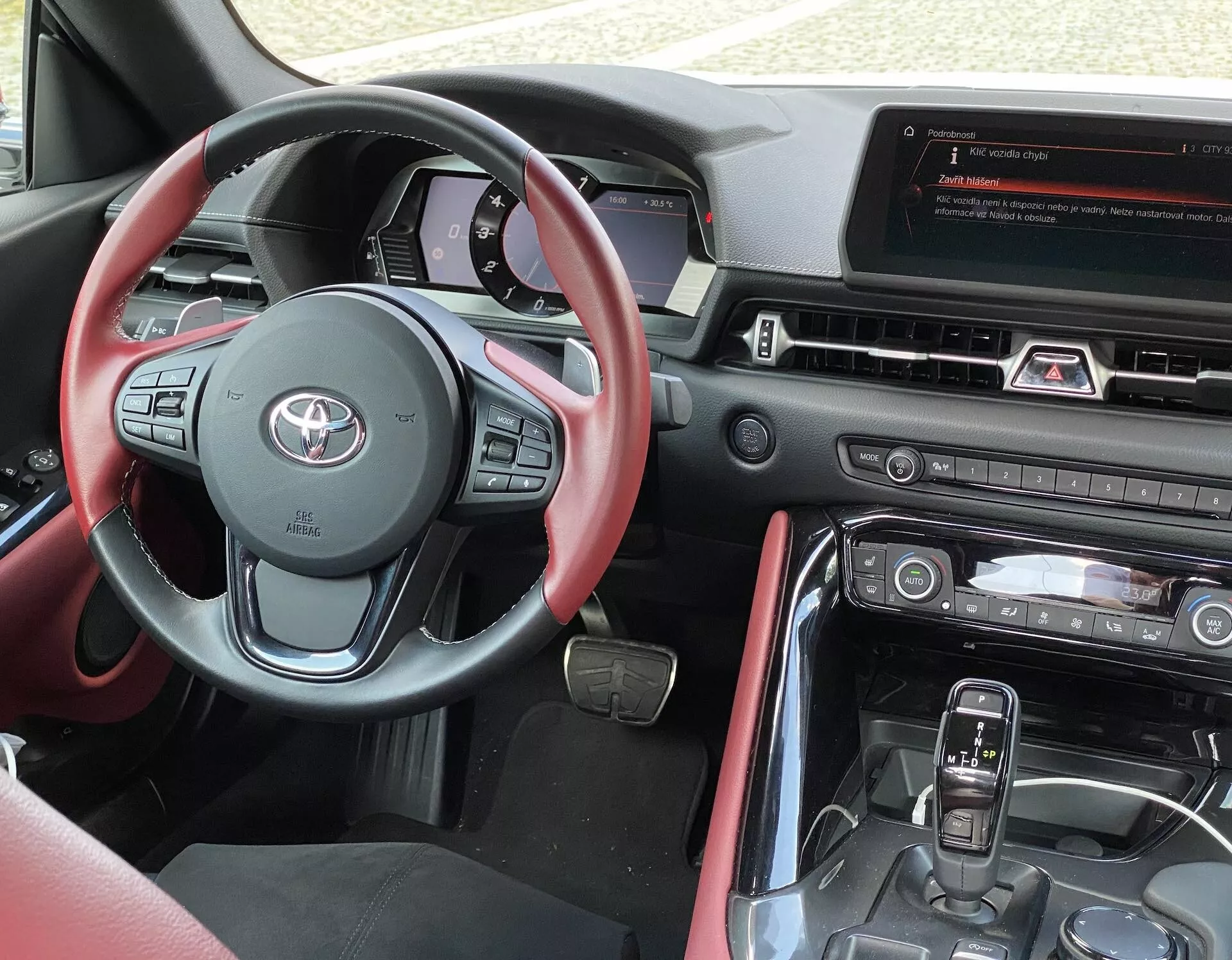 Pohled na interiér z místa řidiče vozu Toyota GR Supra 2.0 Pure Automatic 2019, který ukazuje moderní multifunkční volant s červeným ozdobným prošitím a různými ovládacími tlačítky, digitální přístrojovou desku za volantem a středový panel s velkým displejem informačního systému. Pod displejem jsou ovládací prvky HVAC a středová konzola zahrnuje automatickou řadicí páku a další ovládací tlačítka vozidla. Interiér je navržen ve dvoubarevném schématu s koženými nebo podobnými materiály a sladěnými červenými akcenty.