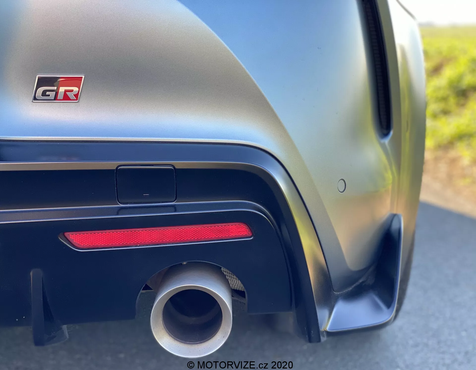 Detailní boční perspektiva zadní čtvrti automobilu Toyota GR Supra 2.0 Pure Automatic 2019, kde se nárazník setkává s karoserií. Detailní pohled zdůrazňuje oblouk kola, velkou kruhovou výfukovou trubku, vodorovný červený odrazka, černý aerodynamický lem a metalický lak měnící se z tmavých na světlé odstíny podél agresivních linií karoserie.