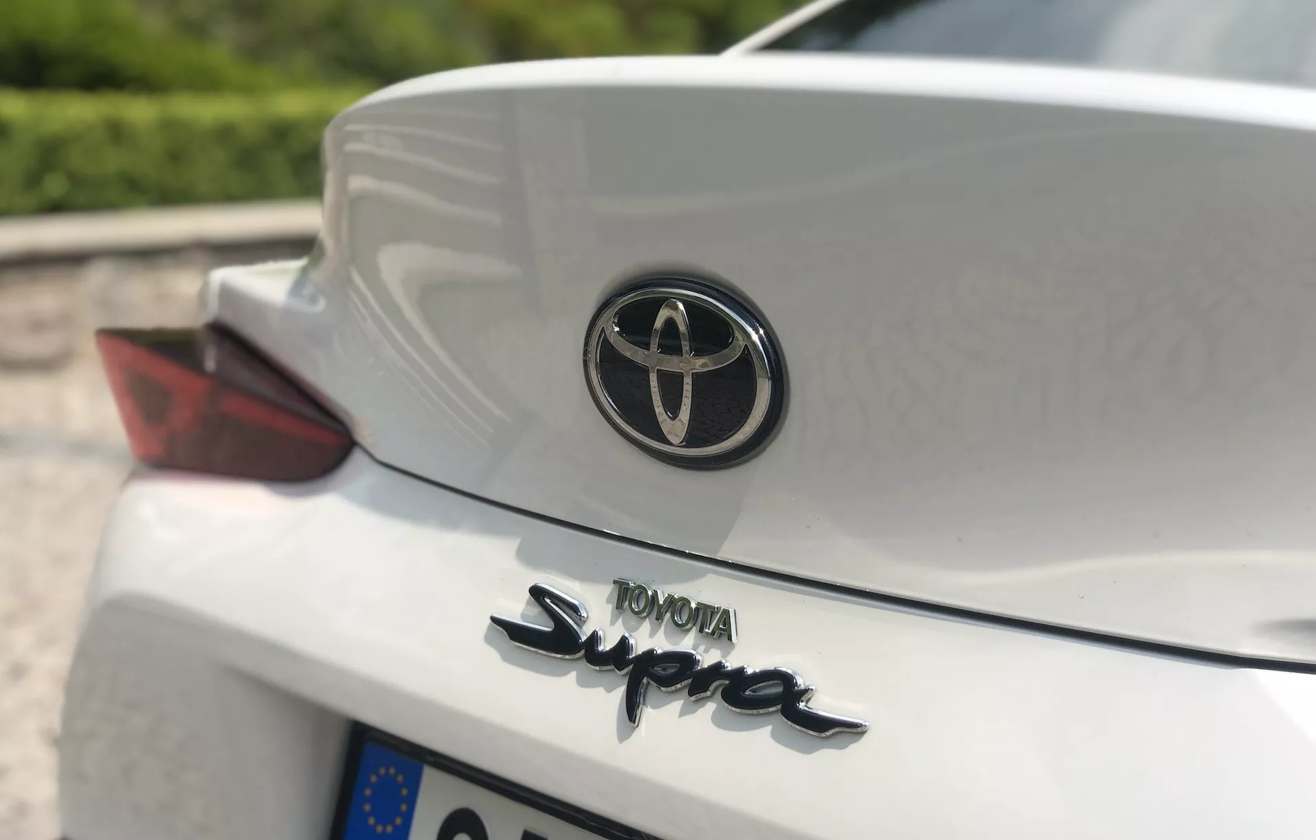 Detailní pohled na zadní část automobilu Toyota GR Supra 2.0 Pure Automatic z roku 2019, snímán z nízkého až středního úhlu, zaměřený na emblém Toyota a výrazný název 'Supra' na bílém víku kufru. Viditelná je i část pasažérského zadního světla s červenou čočkou a nápis 'TOYOTA' velkými písmeny. Vestavěný spoiler přidává sportovní design, s rozmazaným pozadím díky malé hloubce ostrosti.
