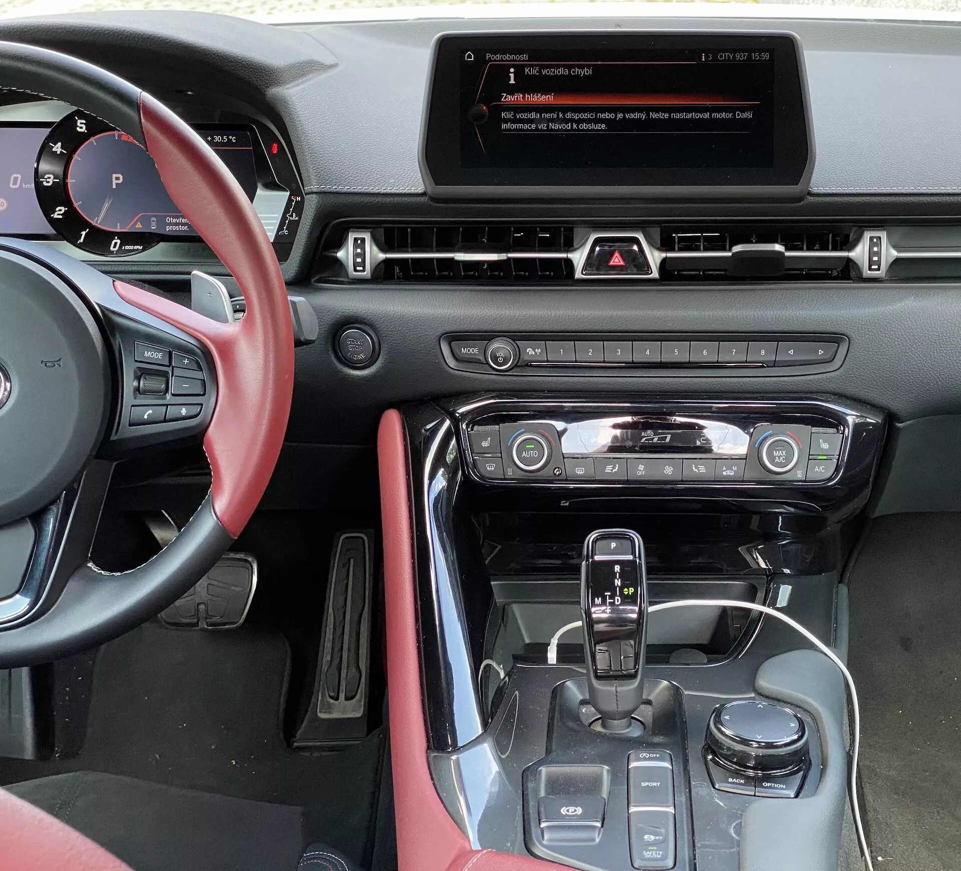 Pohled z místa řidiče v Toyotě GR Supra 2.0 Pure Automatic z roku 2019, zobrazující středový panel, infotainment displej, horizontální větrací otvory, panel varovných světel, systém klimatizace s kulatými ovladači, řadicí páku s lesklým černým lemováním, tlačítka pro jízdní režimy a červené kožené obložení na dveřích řidiče.