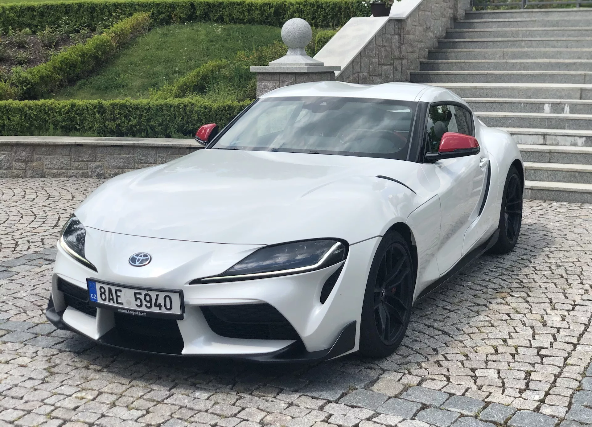Obrázek zobrazuje bílý Toyota Supra 2019 (toyota-gr-supra-2-0-pure-automatic) z předního tříčtvrtečního úhlu. Jasně jsou viditelné nízký a široký postoj auta, ostrá úhlová světla a černé vícedílné hliníkové disky. Fotografie zdůrazňuje elegantní design, aerodynamické kontury a výkonnostní estetiku. Sportovní auto je zaparkováno na dlažbě s obrubníkem a schodištěm v pozadí, což naznačuje městské nebo obytné prostředí.