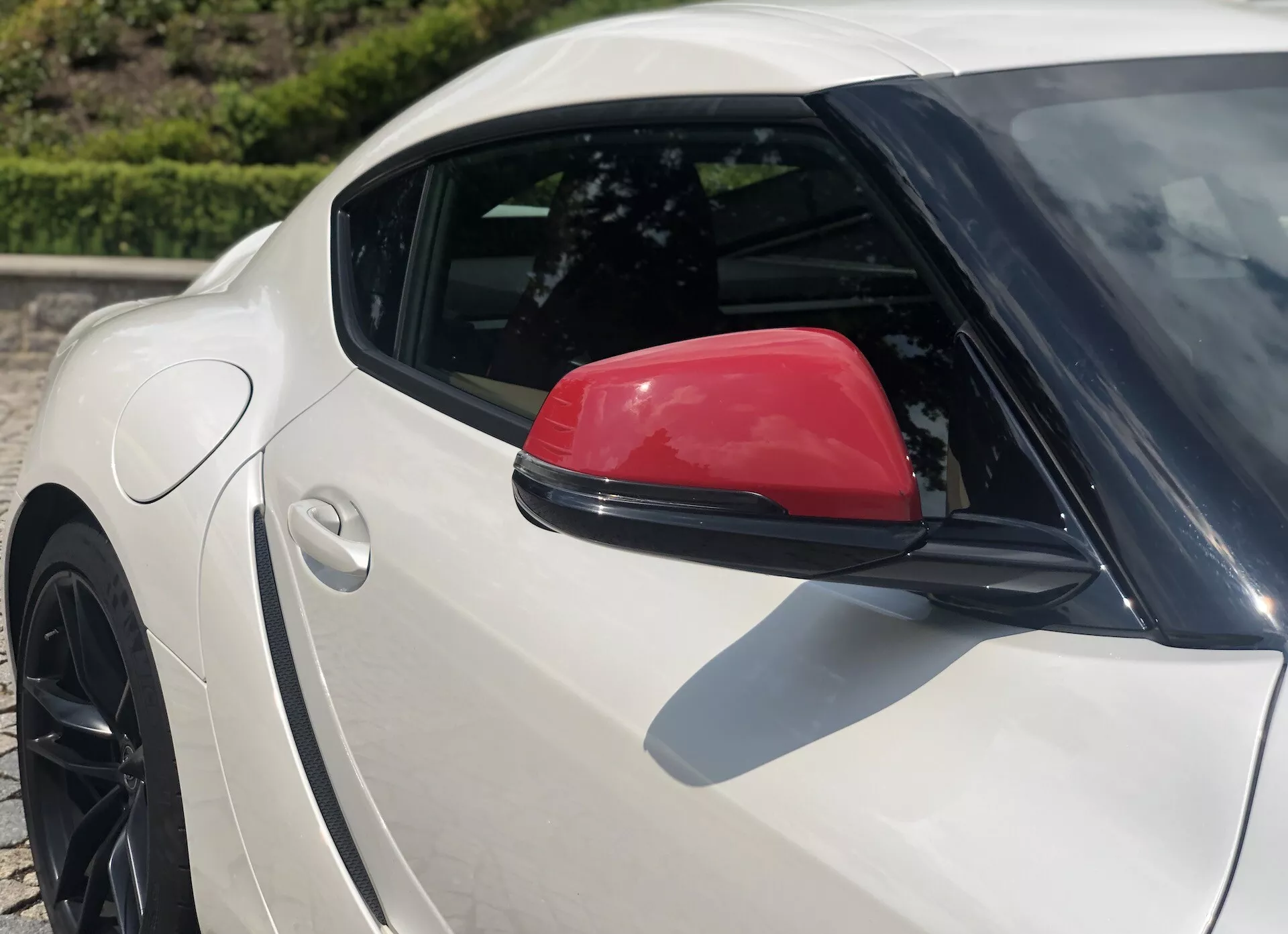 Detailní boční pohled na stříbrnou Toyota GR Supra 2.0 Pure Automatic z roku 2019, zdůrazňující zadní kvartál s charakteristickým červeným bočním zrcátkem a částečně viditelným zadním černým vícepaprskovým litým kolem s nízkoprofilovou pneumatikou; pohled z vyššího úhlu zdůrazňuje křivku zadního blatníku a svažující se střešní linii, situován v dobře osvětleném venkovním prostředí.