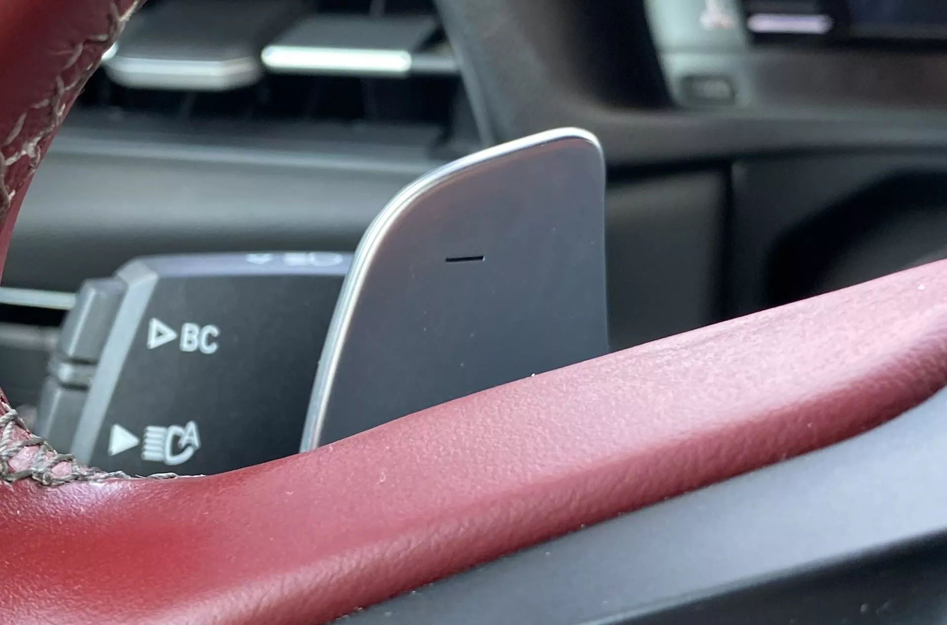 Zblízka z pohledu řidiče Toyota GR Supra 2.0 Pure 2019 automat. Obrázek zdůrazňuje detaily jako červeně potažený volant, částečně viditelné ovládací páčky na sloupku, pádla na řazení a středový držák na mobilní telefon. Malá hloubka ostrosti zdůrazňuje držák na telefon jako centrální prvek, což naznačuje kombinaci sportovních a moderních prvků interiéru.