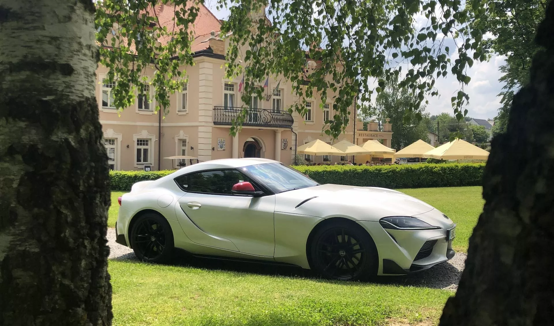 Obrázek zobrazuje Toyota GR Supra 2019 2.0 Pure Automatic, zaparkovanou mezi dvěma stromy, které vytvářejí komponovaný a vyvážený rám. Auto je umístěno na pečlivě upraveném trávníku před elegantním sídlem s žaluziemi. Tento boční pohled zachycuje auto z mírně předního bodu, zdůrazňuje přední kolo a ukazuje hladké designové linie vozu. Jasně bílý kupé má černá boční zrcátka, černé vícekřídlé ráfky a lehce tónovaná okna. Má také dlouhou svažující se kapotu, elegantní aerodynamický profil a malý zadní spoiler na kufru, což naznačuje sportovní charakter vozu.