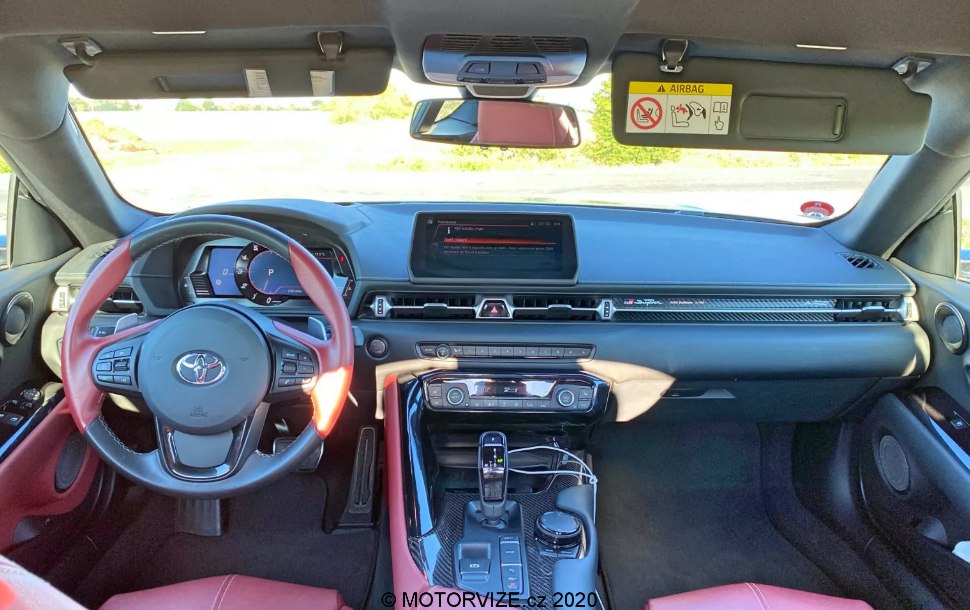 Interiérový snímek Toyota Supra 2019, pohled ze zadního sedadla, ukazuje tříramenný volant s ovladači, vrstvenou palubní desku s centrálním dotykovým infotaimentem, úzký větrací otvor, tlačítka klimatizace a středovou konzoli s automatickou řadicí pákou, elektronickou parkovací brzdou a držáky na nápoje. Červený a černý interiér s částečně viditelnými sportovními sedadly a středovou opěrkou.