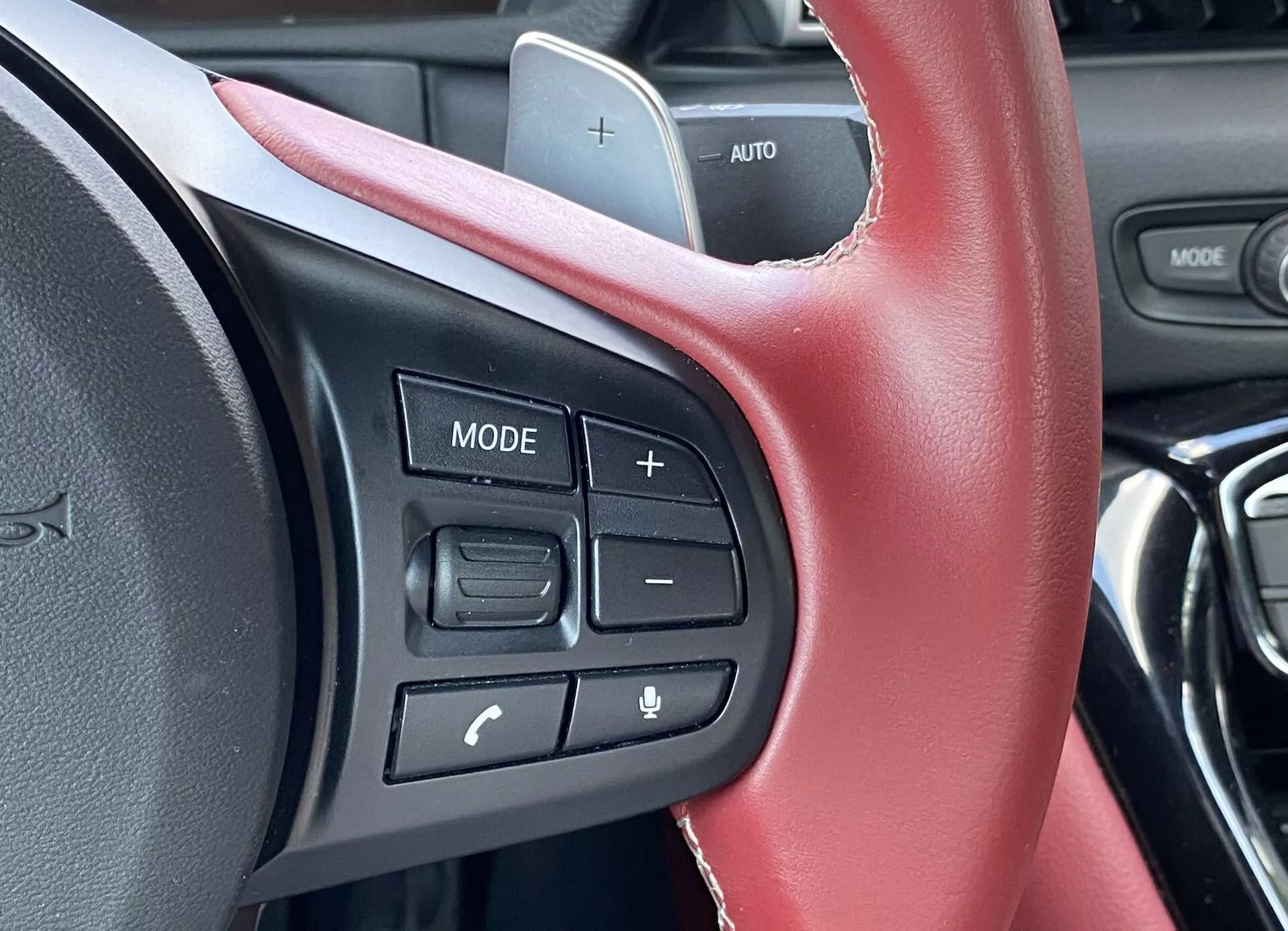 Detailní pohled na volant vozu Toyota Supra 2019 (GR Supra 2.0 Pure Automatic) se zaměřením na ovládací prvky na volantu z pohledu řidiče. Kožený volant s viditelným červeným prošitím naznačuje sportovní interiér. Hmatové tlačítka na levé straně zahrnují tlačítko 'MODE', ovládání hlasitosti označená '+' a '-', a dvě tlačítka pro ovládání telefonu a hlasové příkazy, s nápisem 'AUTO' odkazujícím na tempomat. Design palubní desky je moderní, ale je mimo zaostření, a žádná okolní krajina naznačuje, že auto stojí na místě.