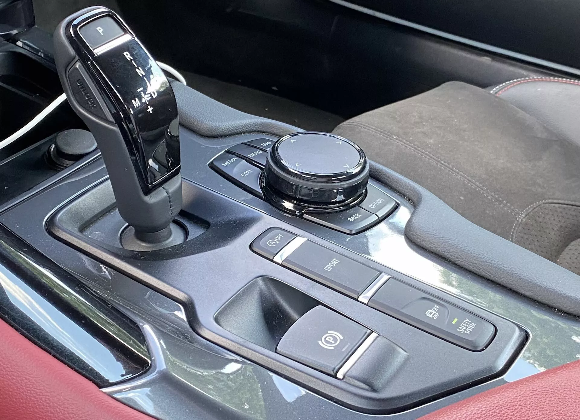 Pohled shora na středovou konzoli interiéru Toyota Supra 2.0 Pure Automatic z roku 2019. Páka řazení je v poloze parkování, obklopená různými ovládacími prvky, včetně otočného ovladače infotainmentu a tlačítek pro dynamiku vozidla a zkratky infotainmentu. Tlačítko start/stop motoru je umístěno prominentně. Konzole obsahuje kovově vypadající akcenty, leštěný kov nebo vysoce lesklý plast a opěrky na ruce z textilu nebo kůže s akcentním prošitím.