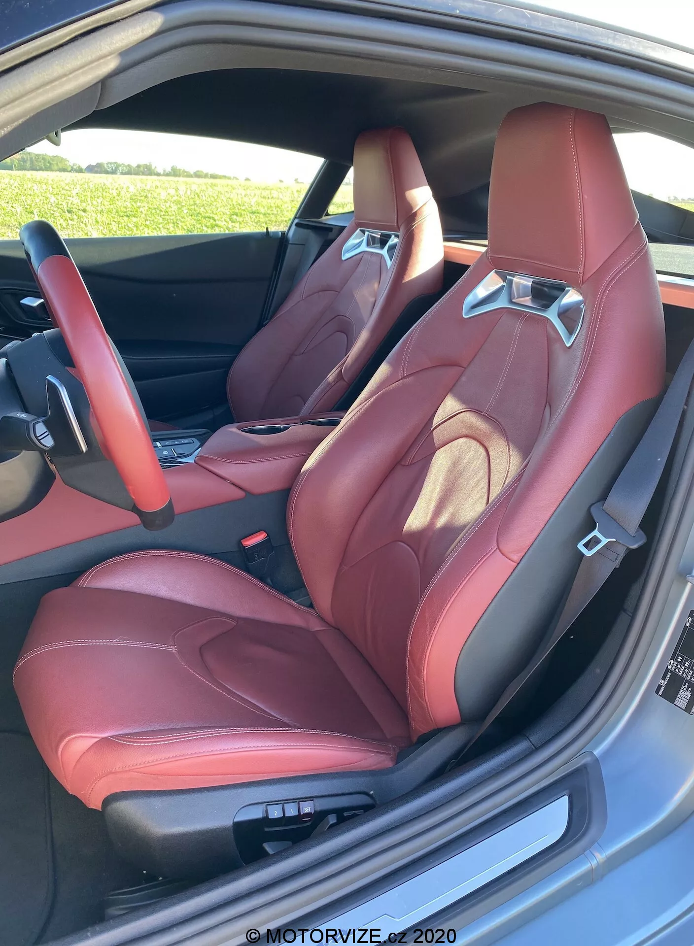 Vysoké rozlišení obrázku sportovního kbelíkového stylu předního sedadla Toyota GR Supra 2019 s červenou koženou čalouněním, výraznými bočními podpěrami, integrovanou opěrkou hlavy a kontrastním švem. Zaznamenáno zvenčí skrz otevřené dveře, odhaluje elektronické ovládání sedadla a částečný pohled na luxusní interiér dveří, čímž zvýrazňuje prémiový a výkonově orientovaný estetický vzhled.